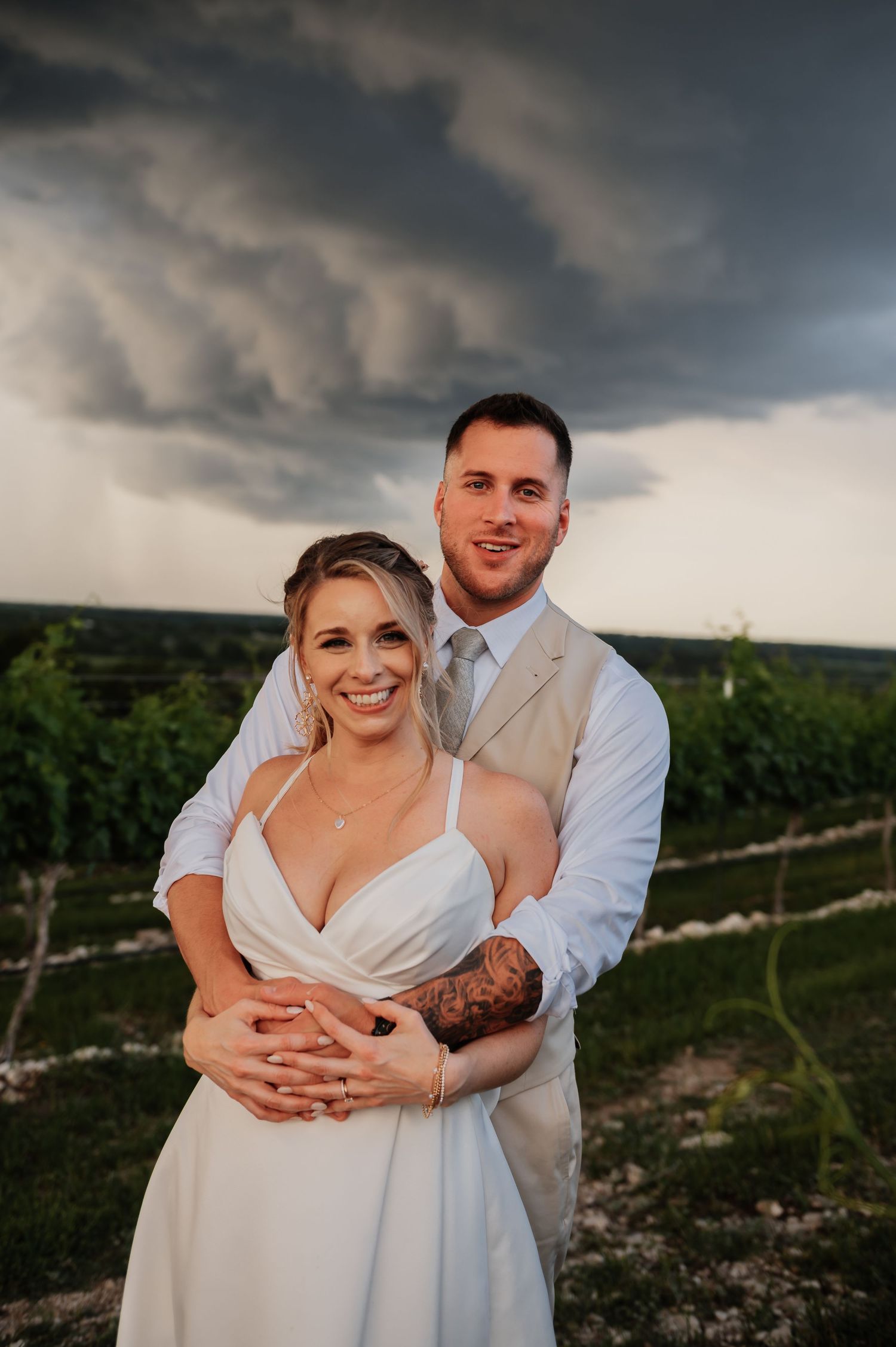 Bride and groom embrace outdoors at vineyard with dramatic stormy sky, groom showing sleeve tattoo.