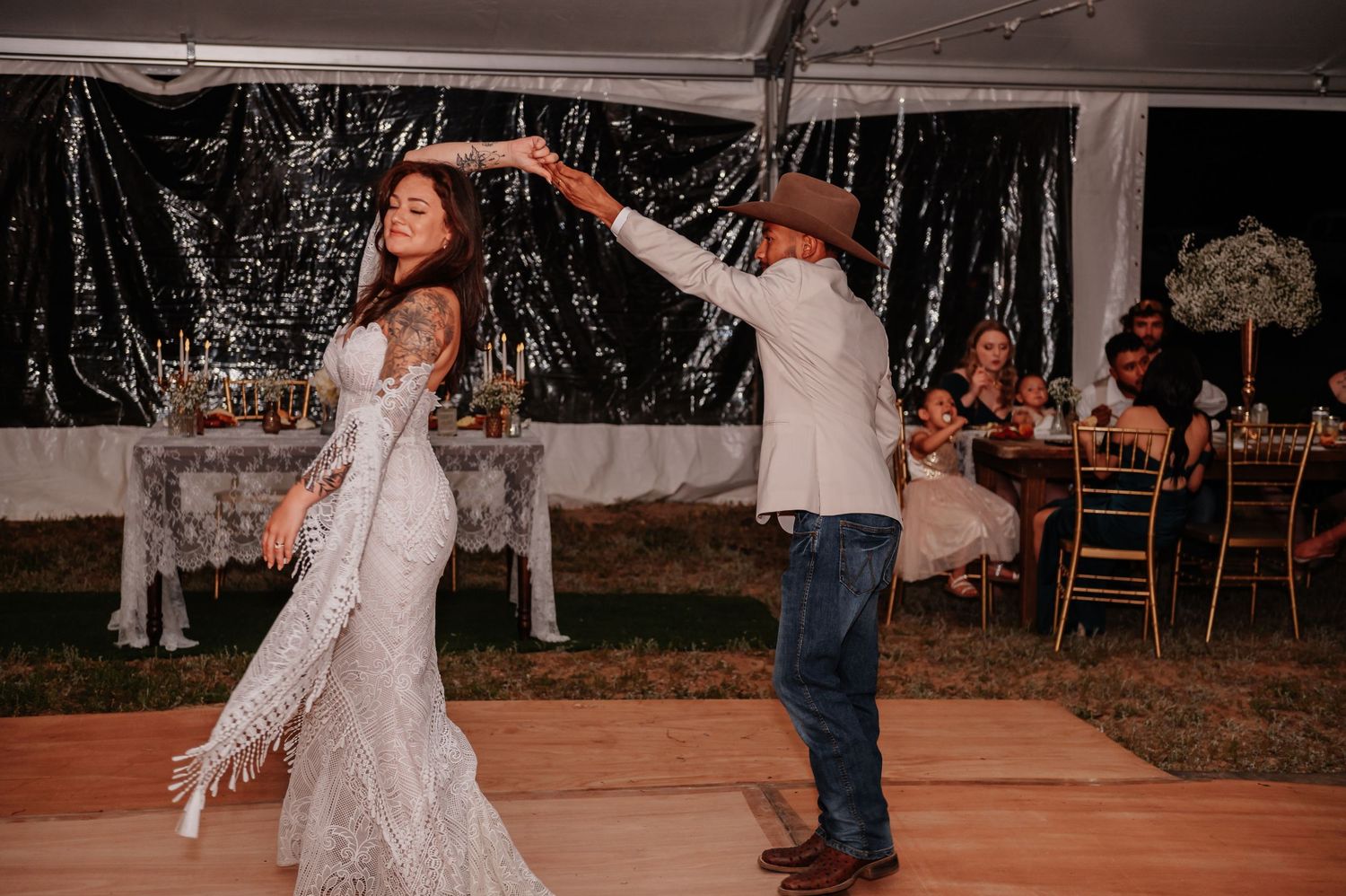 Bride in lace fringe wedding dress twirling with groom in cowboy hat during first dance at outdoor tent reception.