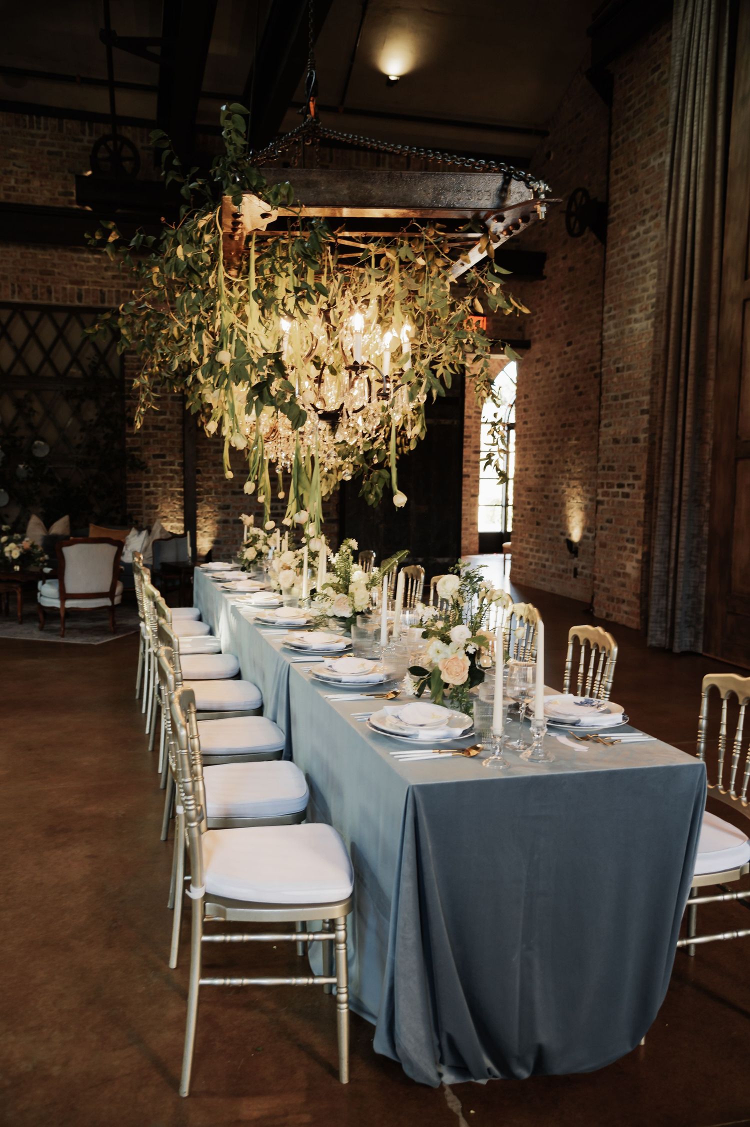 Elegant wedding reception with dusty blue tablecloth, white place settings, and dramatic hanging floral chandelier.