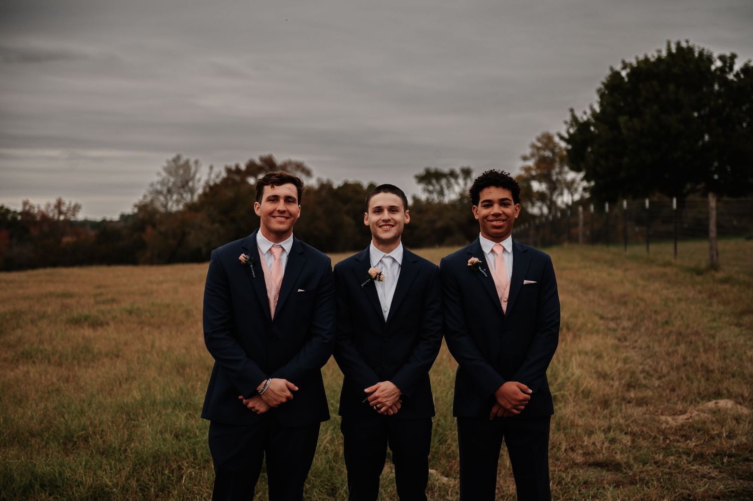 Three groomsmen in navy suits with pink ties smiling outdoors in a rustic field on a cloudy wedding day.