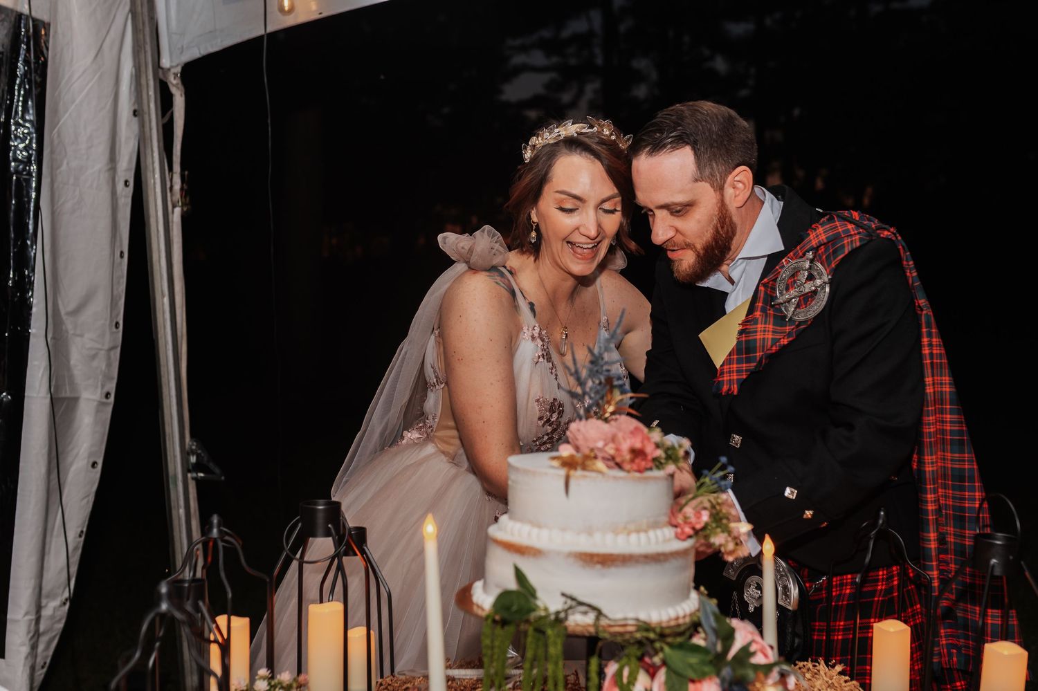 Bride and groom cutting their wedding cake together, groom in a red tartan kilt, surrounded by candles and florals.