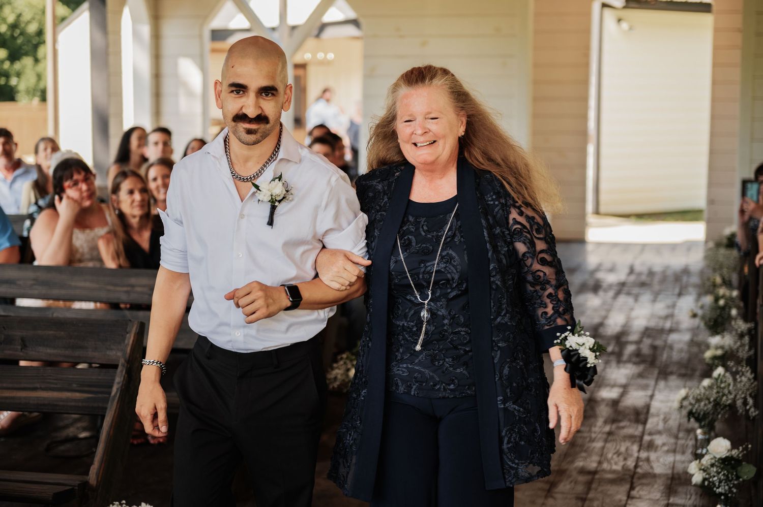 A man in a white shirt with a boutonniere walks arm-in-arm with a smiling woman in navy at a wedding ceremony.