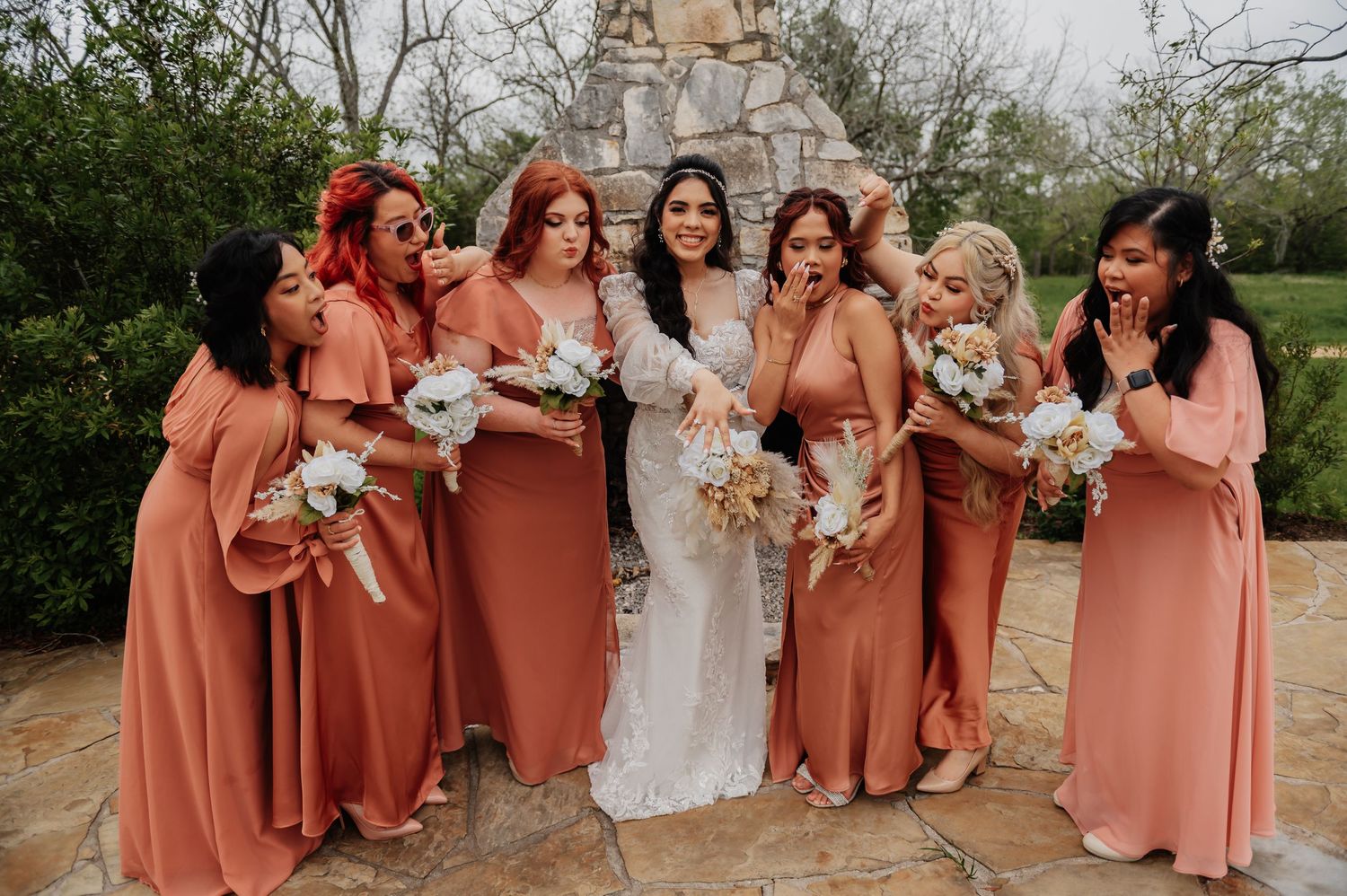 Bride in white lace gown poses with six bridesmaids in coral dresses holding white bouquets outdoors.