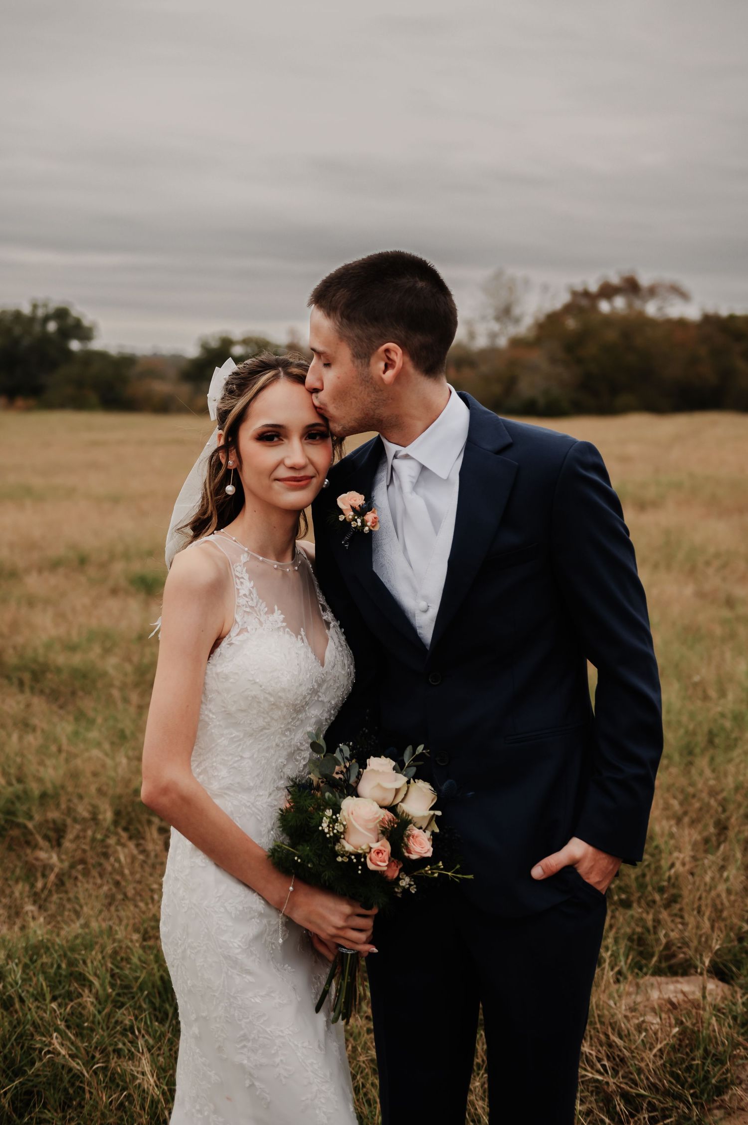 Bride holding pink bouquet smiles softly as groom in navy suit kisses her temple in a meadow.