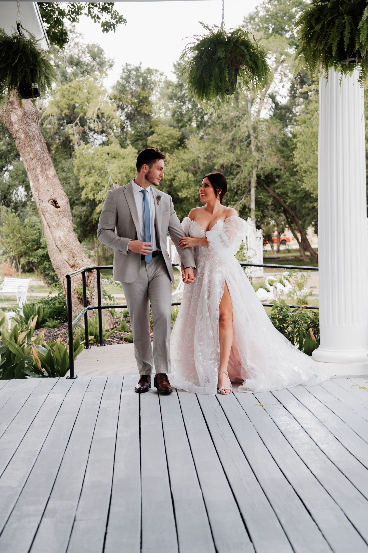 Bride and groom walking on a white wooden porch surrounded by lush greenery on their wedding day.
