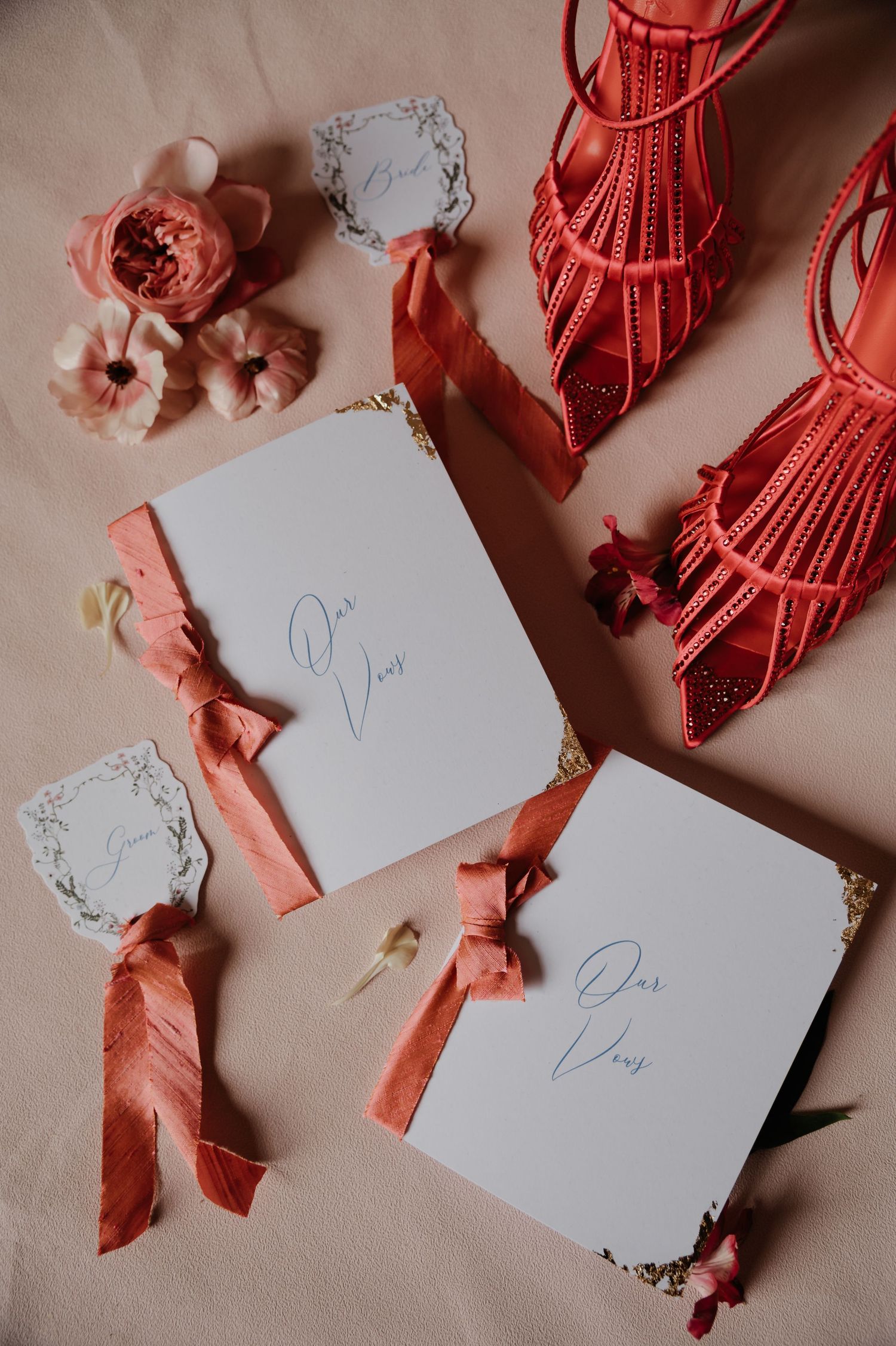Elegant wedding vow books with coral ribbon bows, marble tags, and red strappy heels on a blush pink surface.