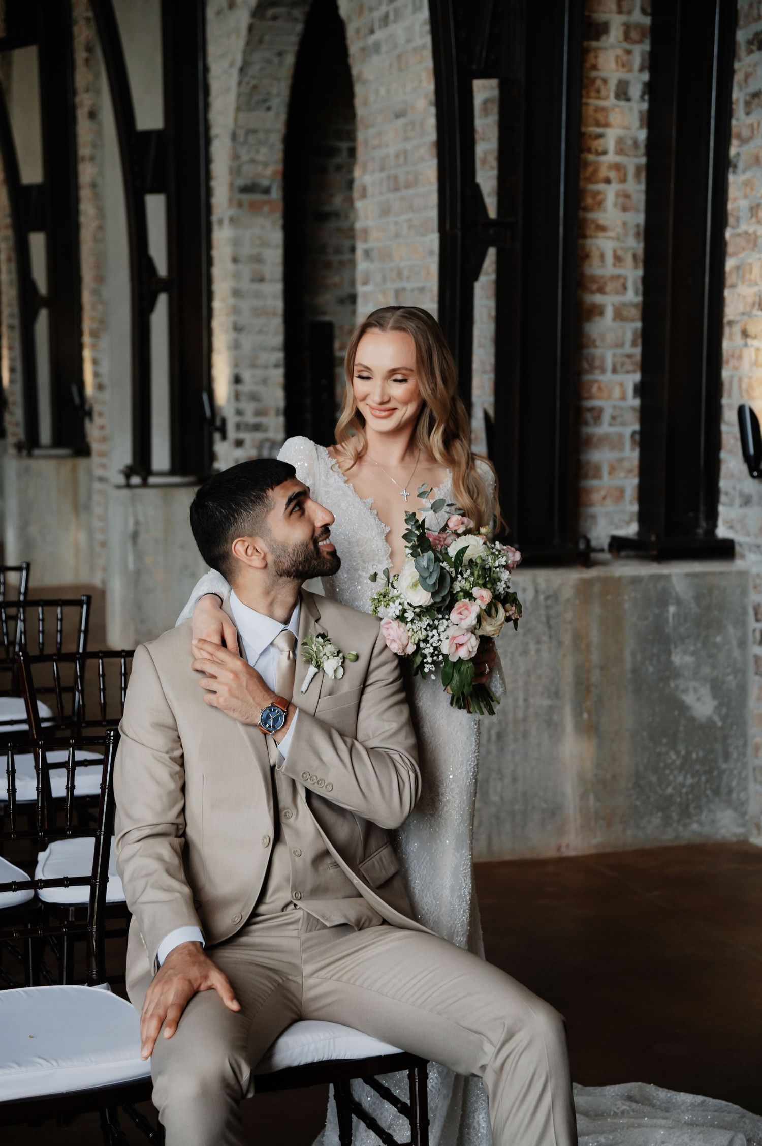 Bride holding pink bouquet stands behind seated groom in tan suit, posing in elegant brick venue.