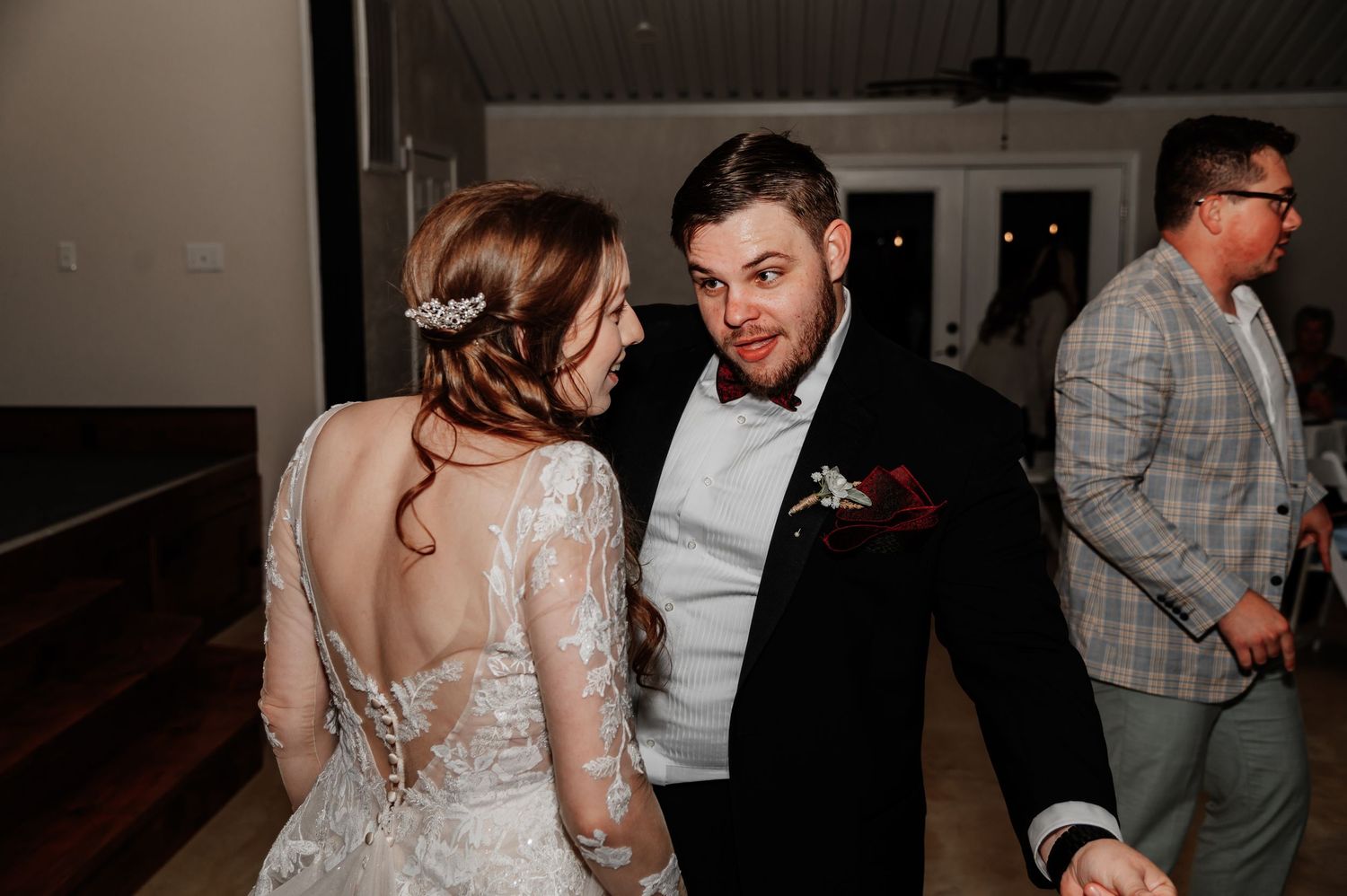 Bride in lace backless gown and groom in black tuxedo share a joyful dance moment at their wedding reception.