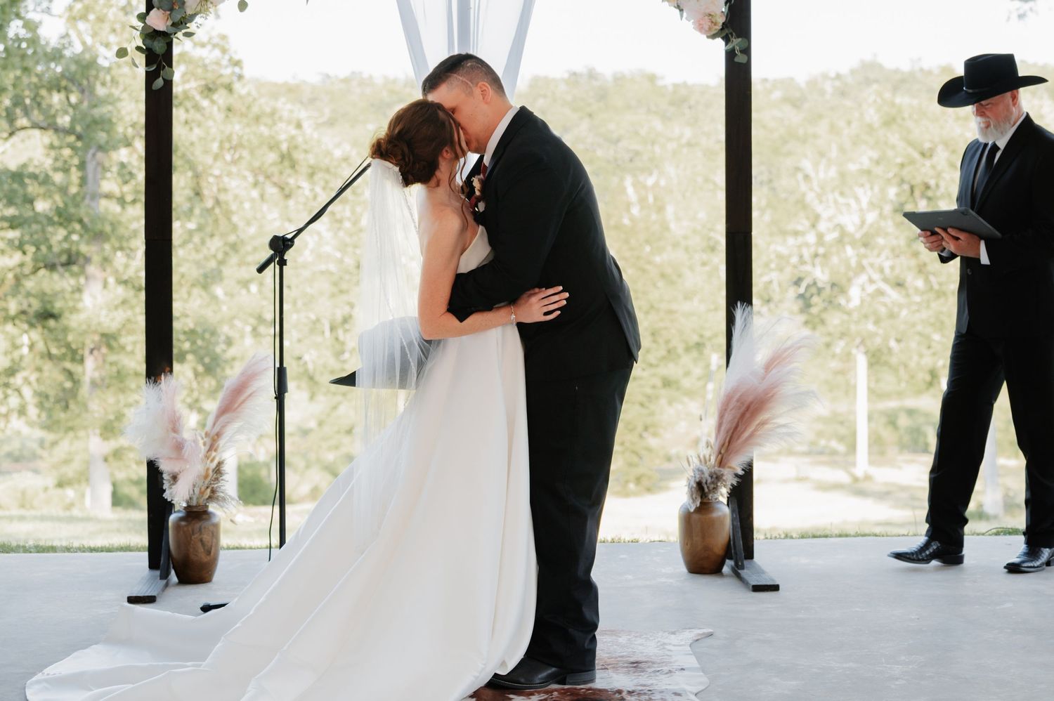 Bride and groom share first kiss at outdoor wedding ceremony with pampas grass decor and officiant nearby.