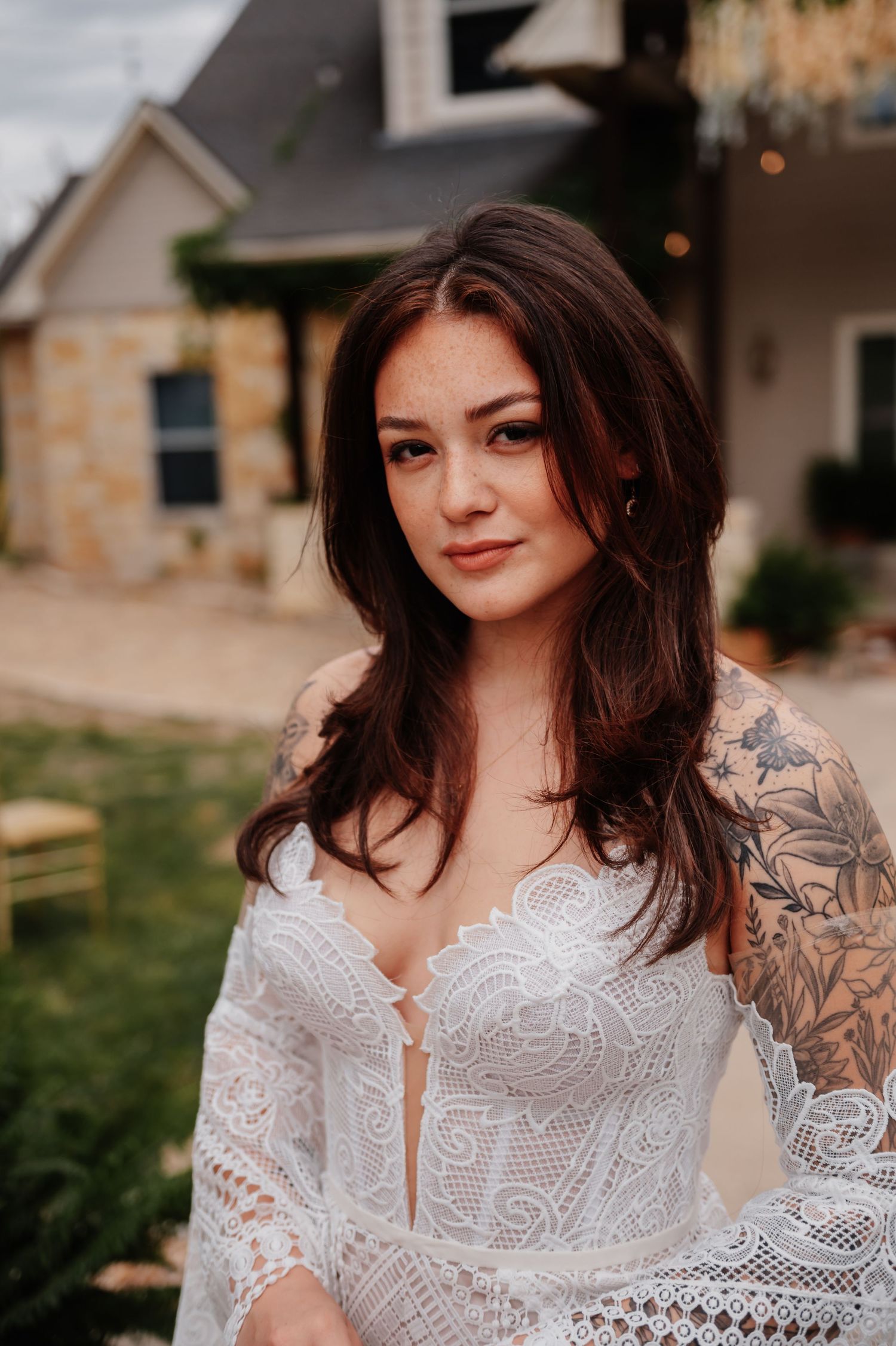 Bride in white lace gown with floral tattoos poses outdoors near stone building.