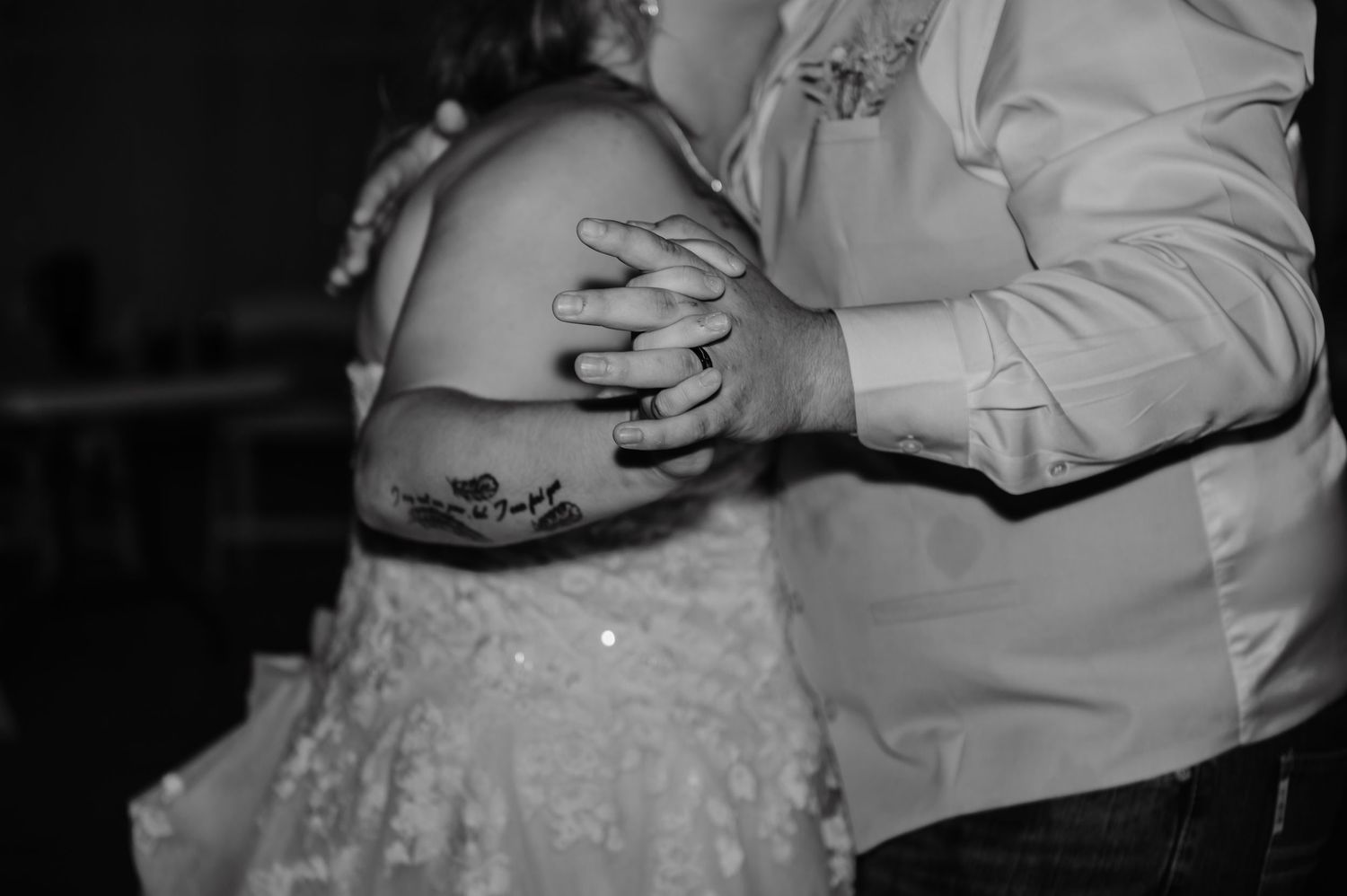 Black and white photo of a couple dancing, hands clasped, bride in lace dress with tattoo visible on arm.