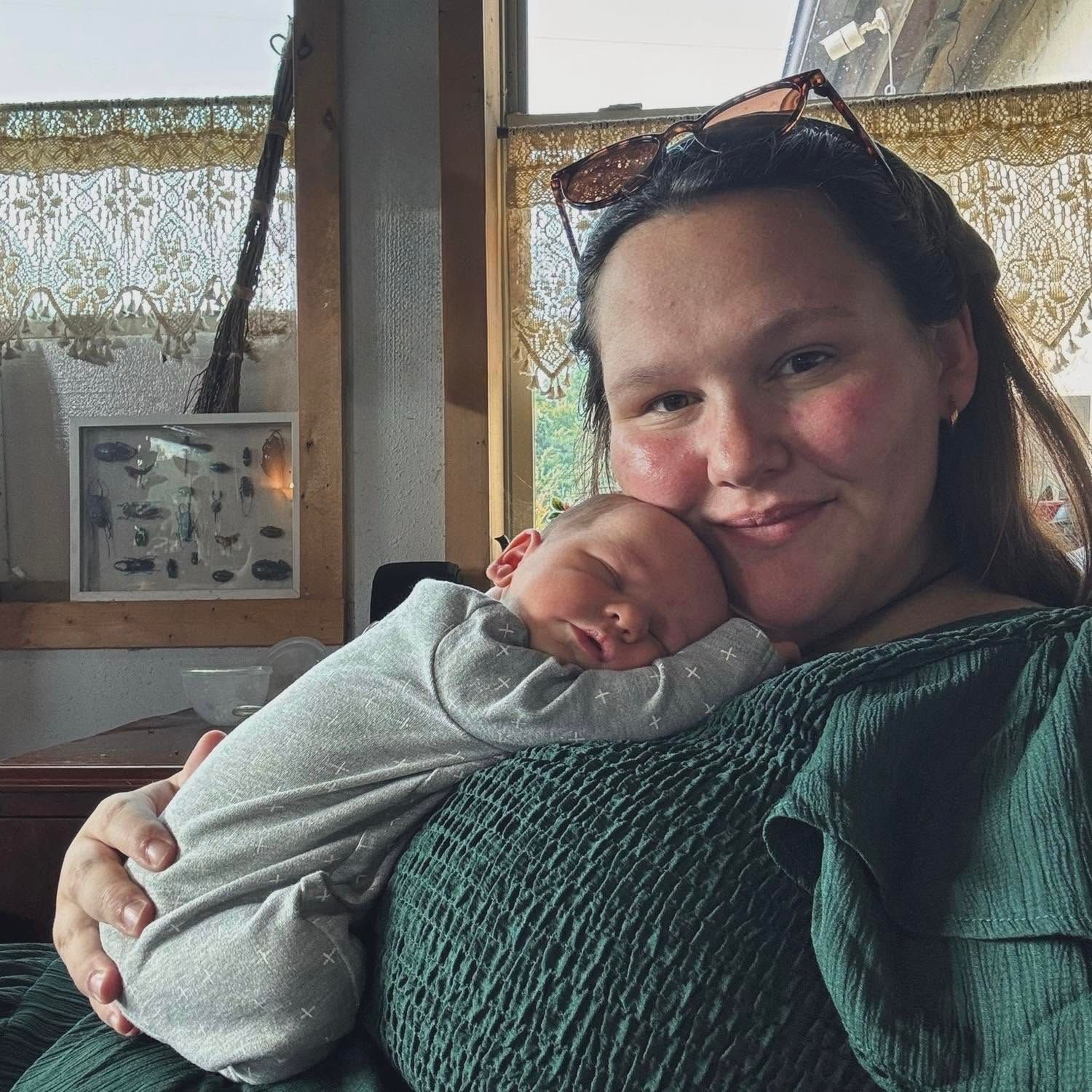A young woman with sunglasses on her head smiles while cradling a sleeping newborn baby indoors.