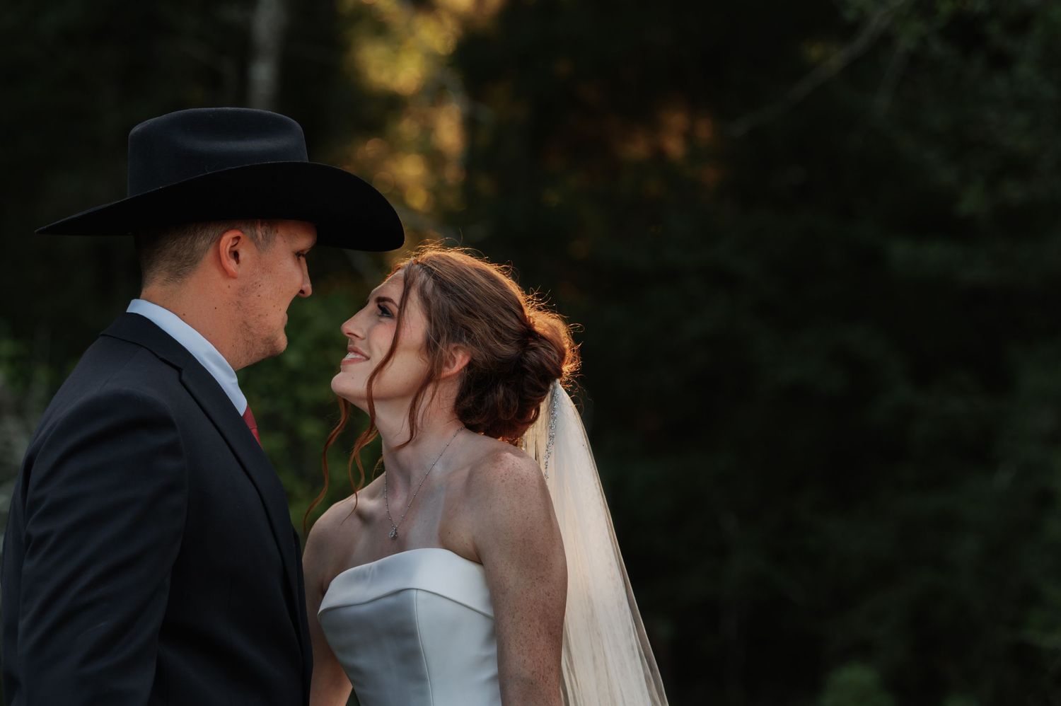 Bride and groom share a romantic moment outdoors, groom wearing a black cowboy hat and suit, bride in a strapless gown with veil.