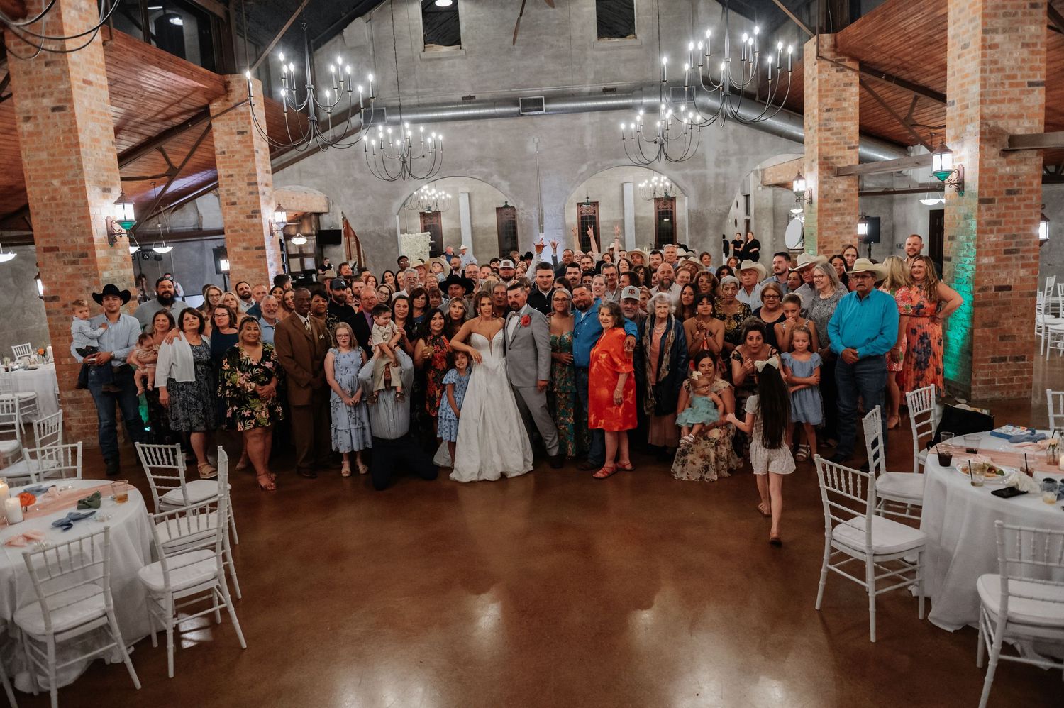 Wedding guests gather for a group photo with bride and groom in a rustic brick venue with string lights.