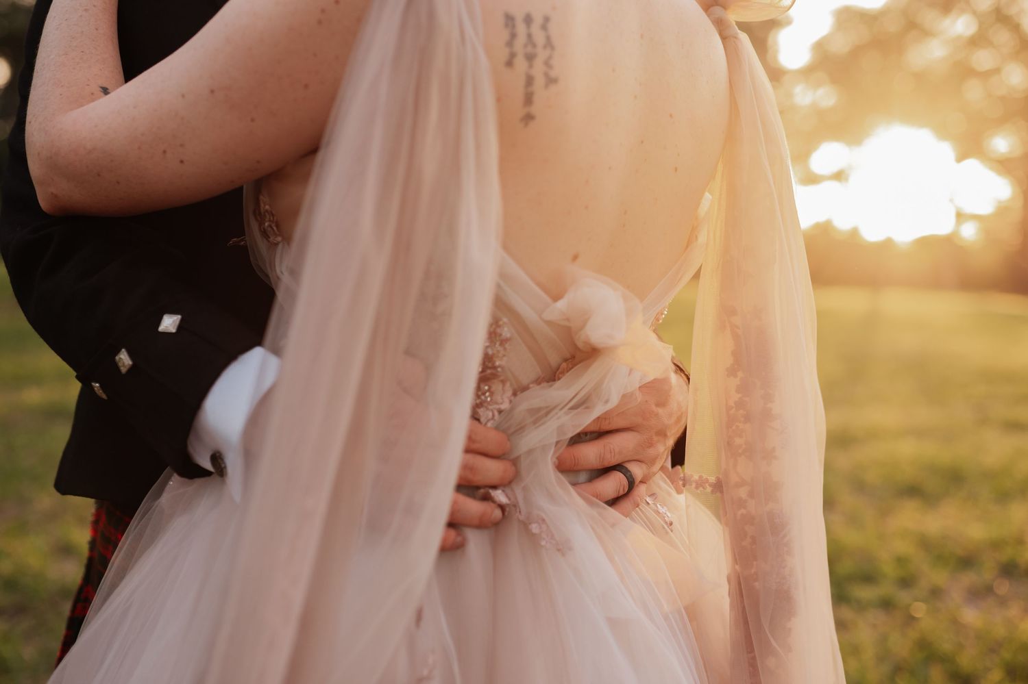 Couple embracing at sunset, bride in blush wedding gown with lace details and veil, groom in dark suit outdoors.