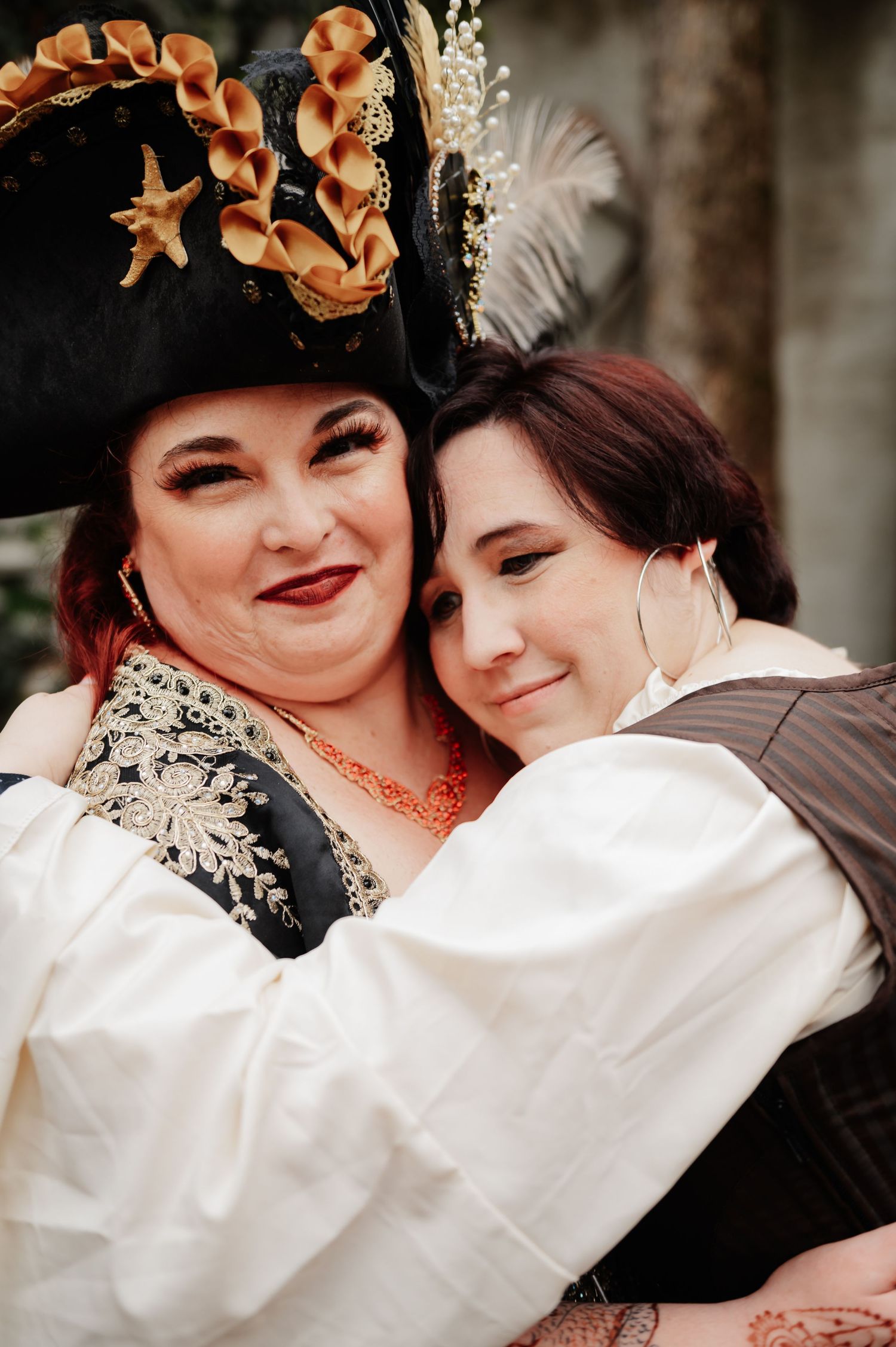 Two women in pirate costumes embrace warmly, one wearing an ornate tricorn hat with flowers and gold embroidery.
