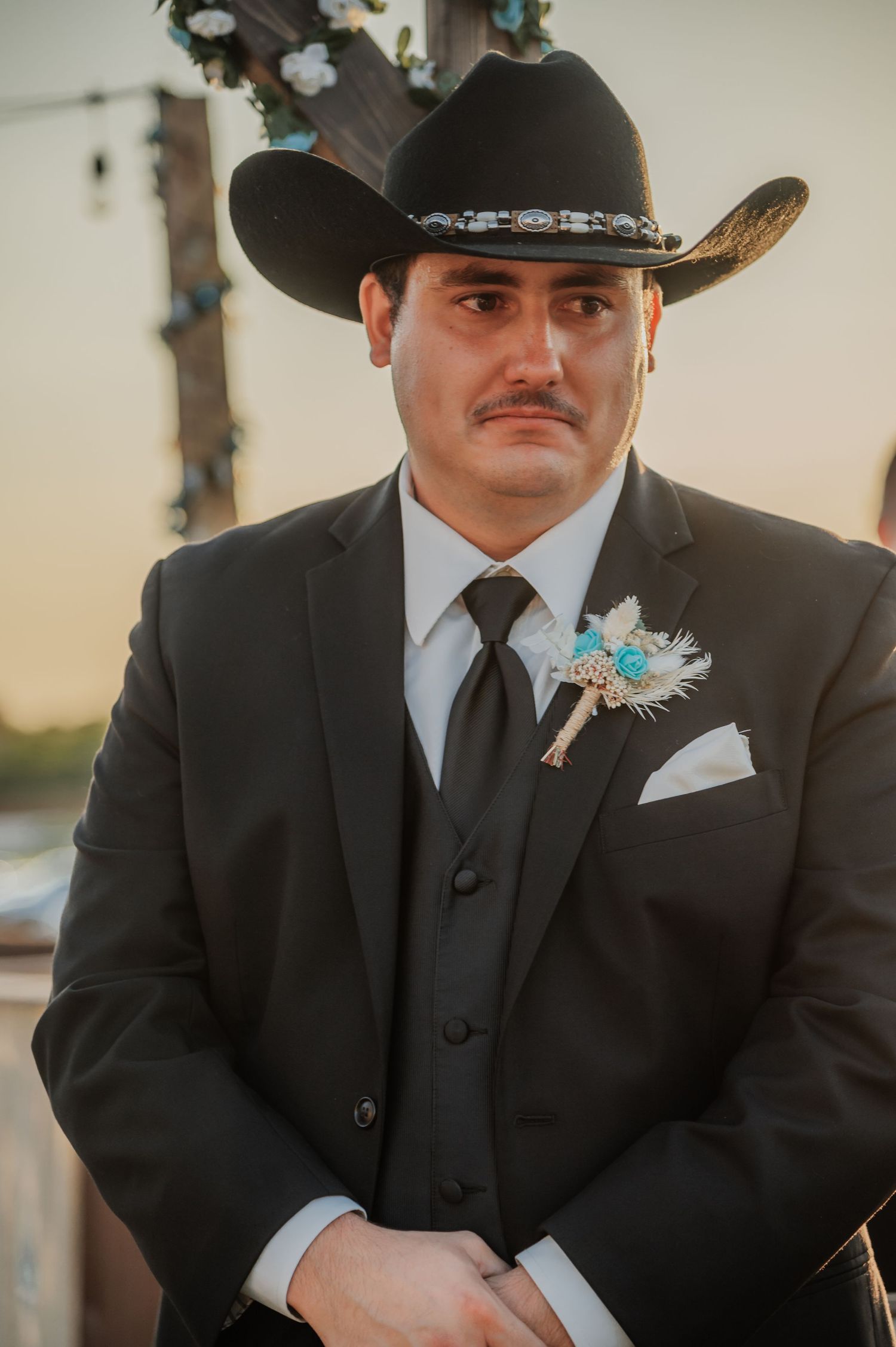 A groom in a black cowboy hat and suit with a turquoise boutonniere stands emotionally at an outdoor wedding ceremony.