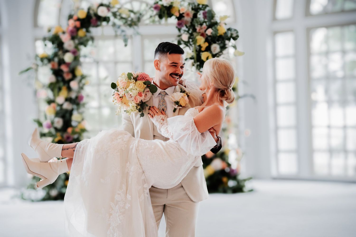 Groom lifting bride in white lace dress, laughing joyfully before a colorful floral arch in bright venue.