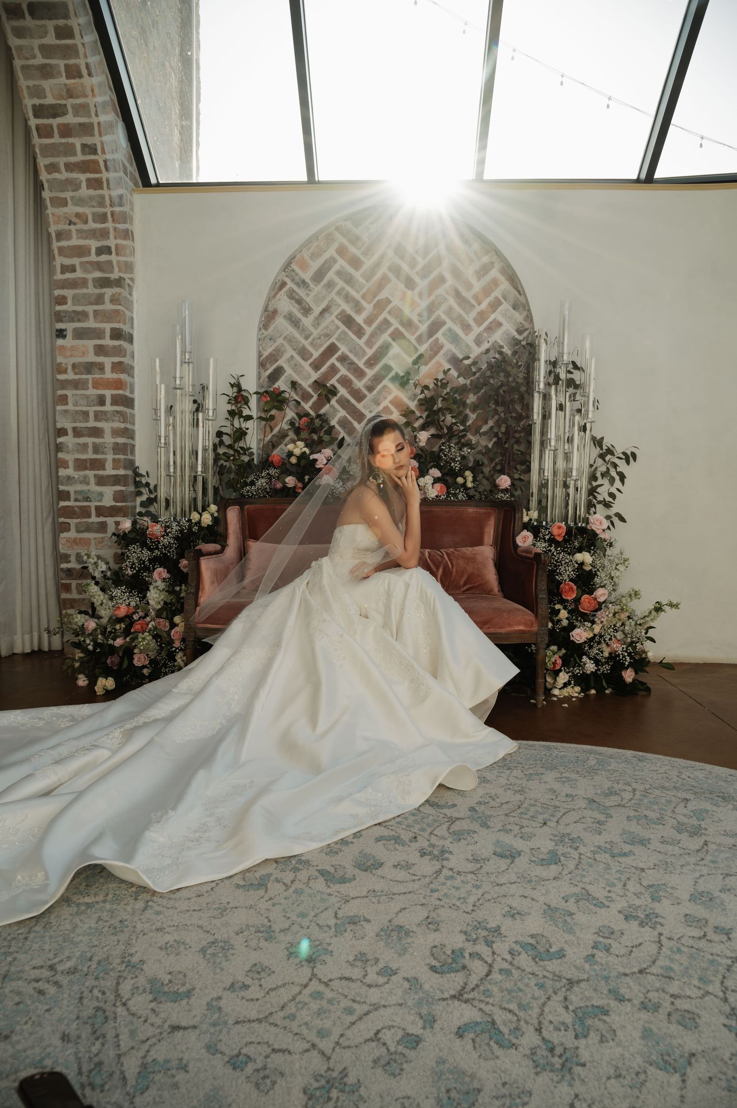 Bride in flowing white gown and veil seated before arched brick altar with candles and floral arrangements.