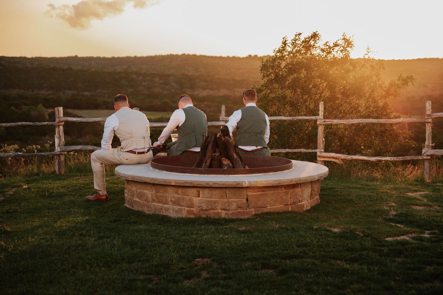 Three men sitting around a stone fire pit at sunset with rustic fence and rolling hills in background.