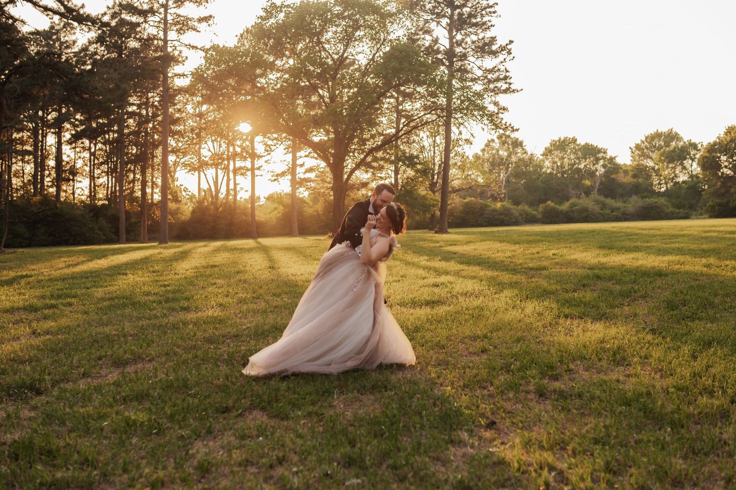 Bride and groom share a romantic dance at golden sunset in a lush green field surrounded by tall trees.
