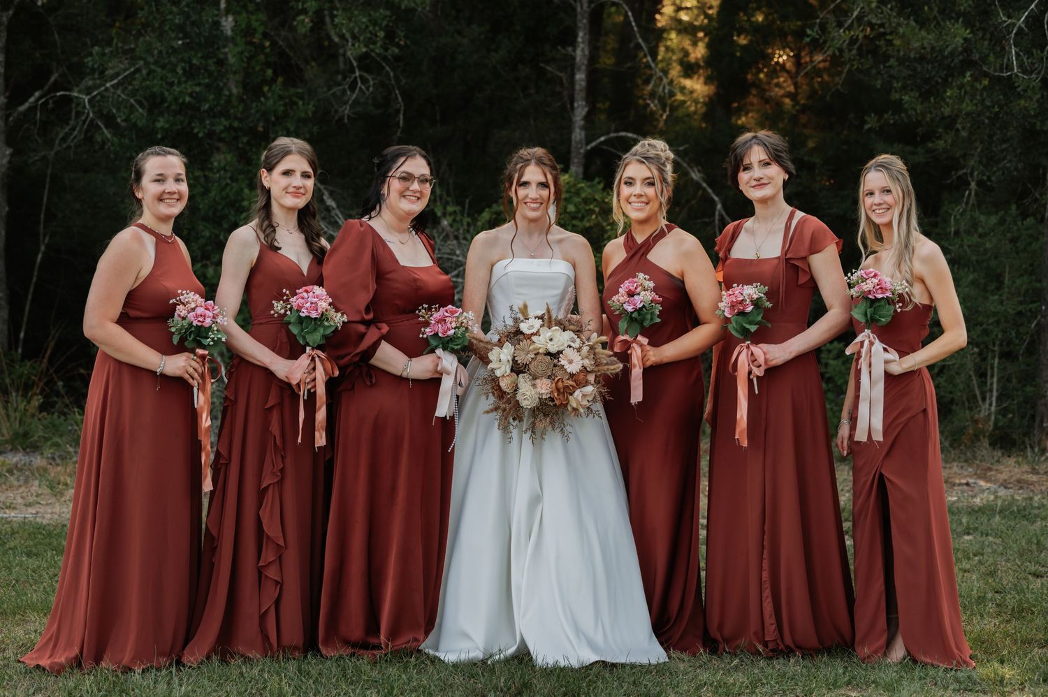Bride in white gown poses with six bridesmaids in rust-colored dresses holding bouquets outdoors.