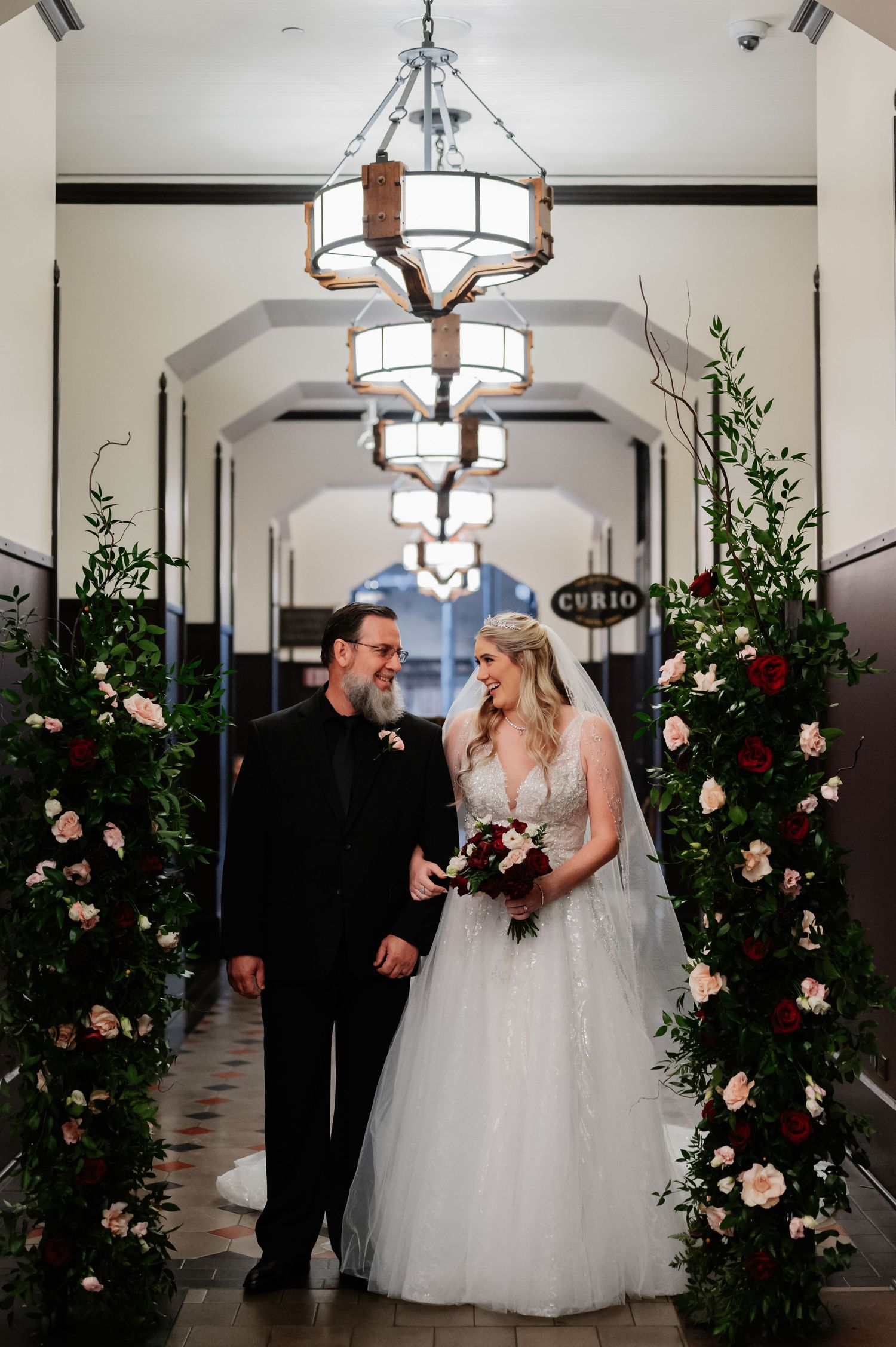 Bride and groom stand beneath floral arches with roses in an elegant hotel corridor with chandeliers.