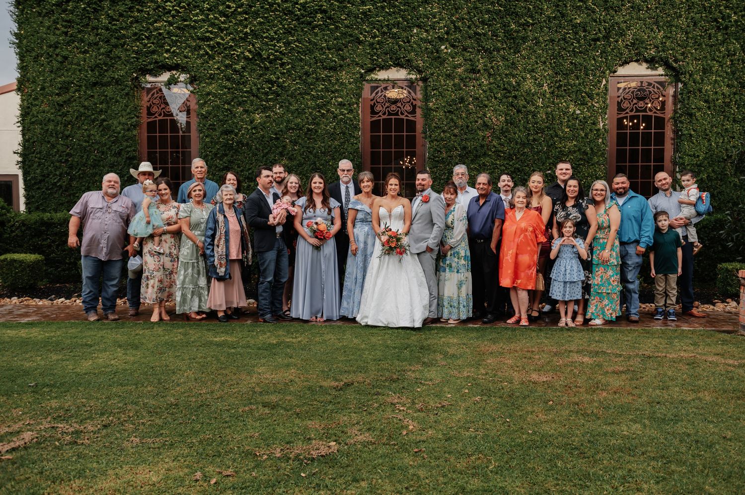 Large wedding group photo with bride and groom surrounded by family outdoors against ivy-covered building wall.