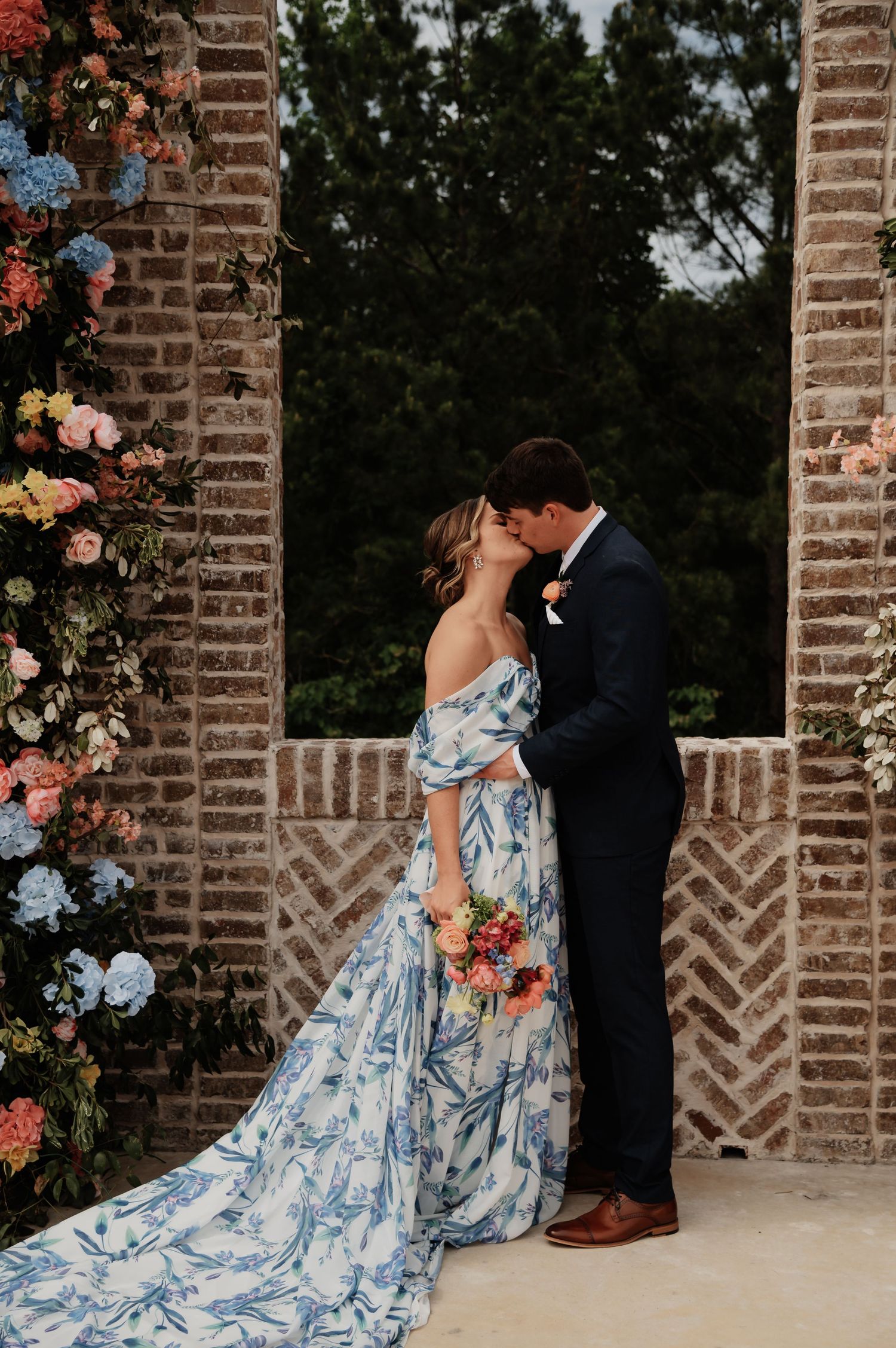 Bride in blue floral off-shoulder gown kisses groom in navy suit at brick arch adorned with colorful flowers.