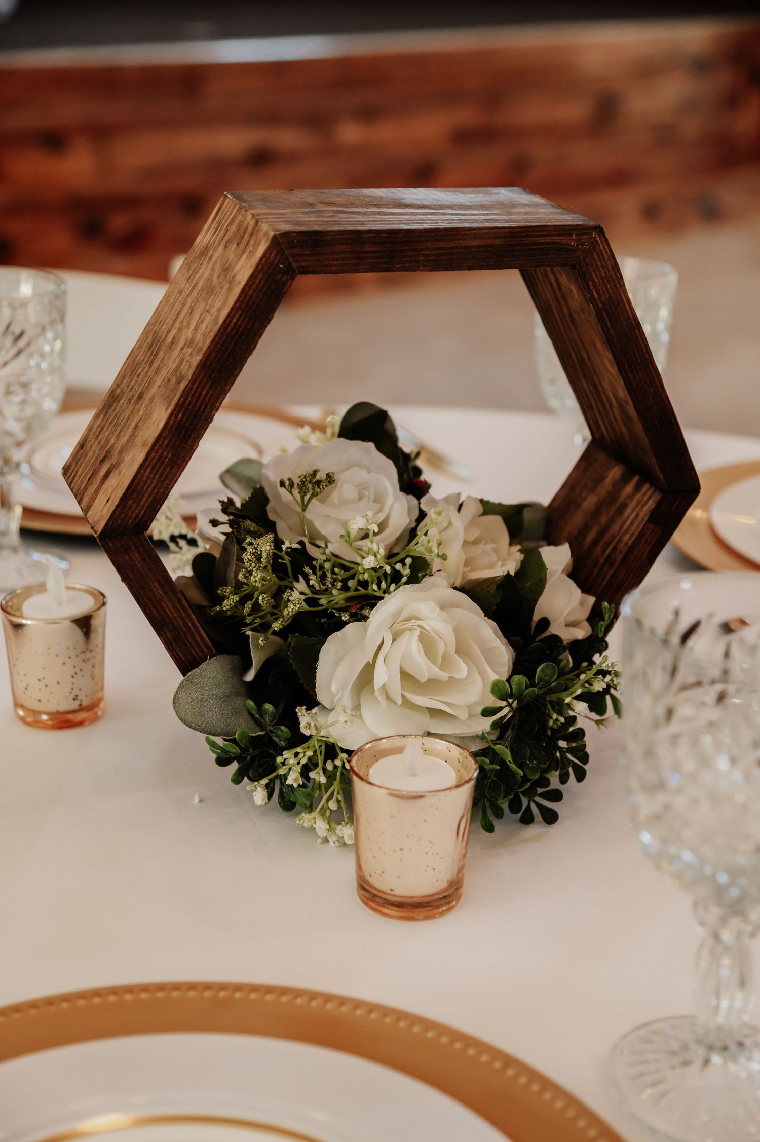 Rustic wooden hexagon centerpiece with white roses, greenery, and gold mercury glass votive candles on elegant table.