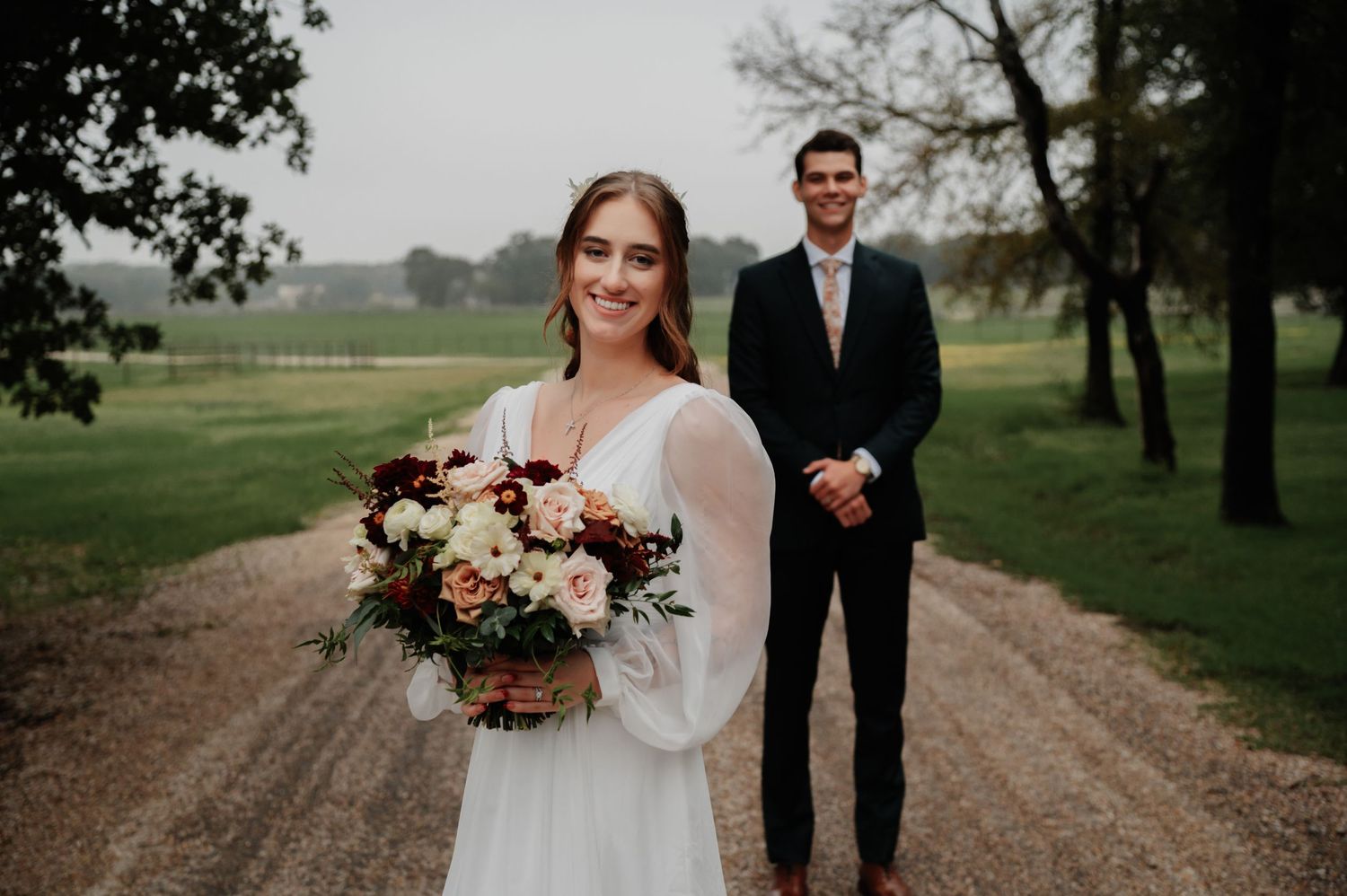 Bride holding floral bouquet on dirt road with groom standing behind her at outdoor wedding venue.