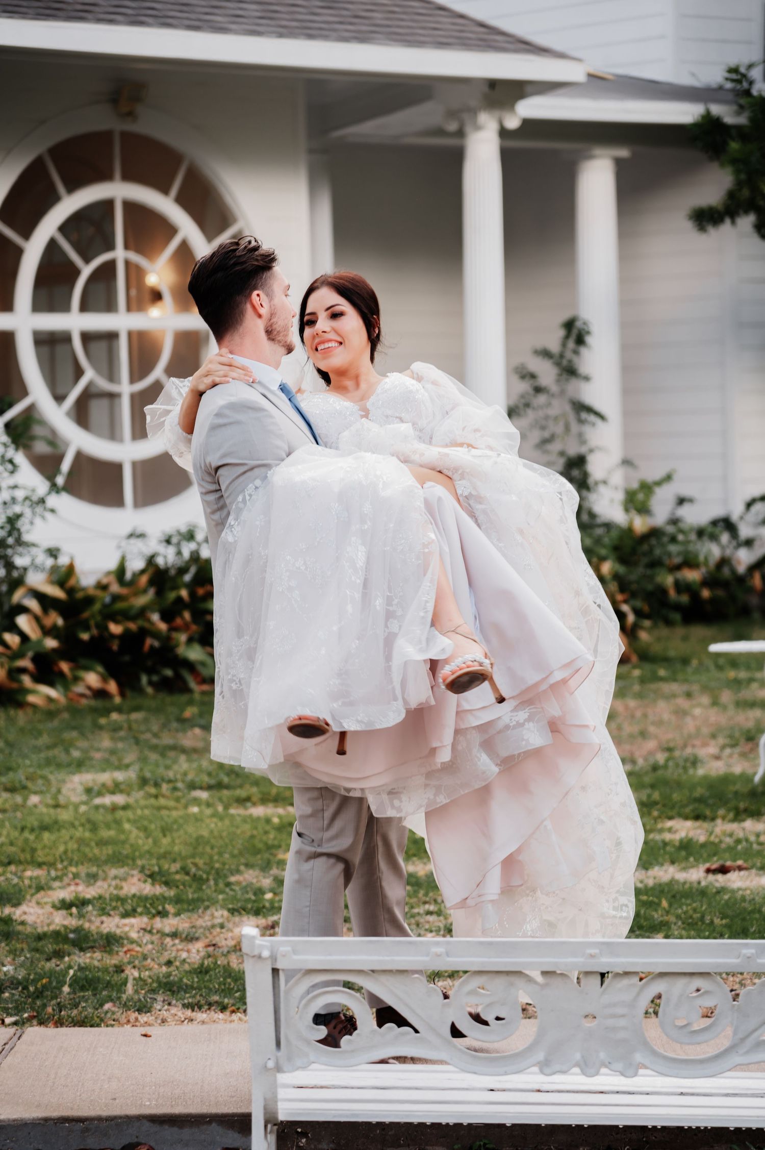 Groom carries bride in white wedding dress outside elegant white venue with manicured lawn.