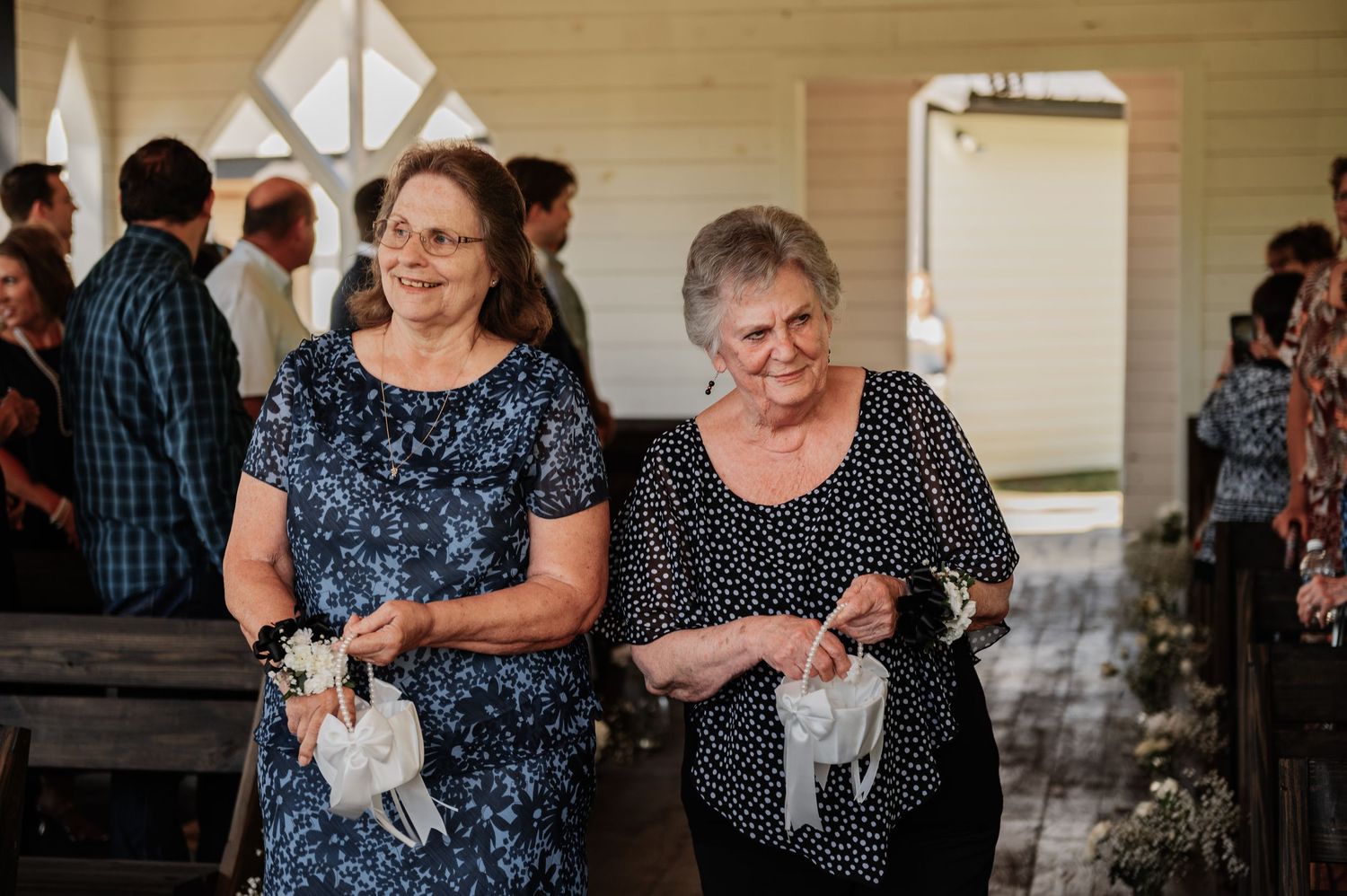 Two elderly women walk down a wedding chapel aisle holding white handkerchiefs during a ceremony.