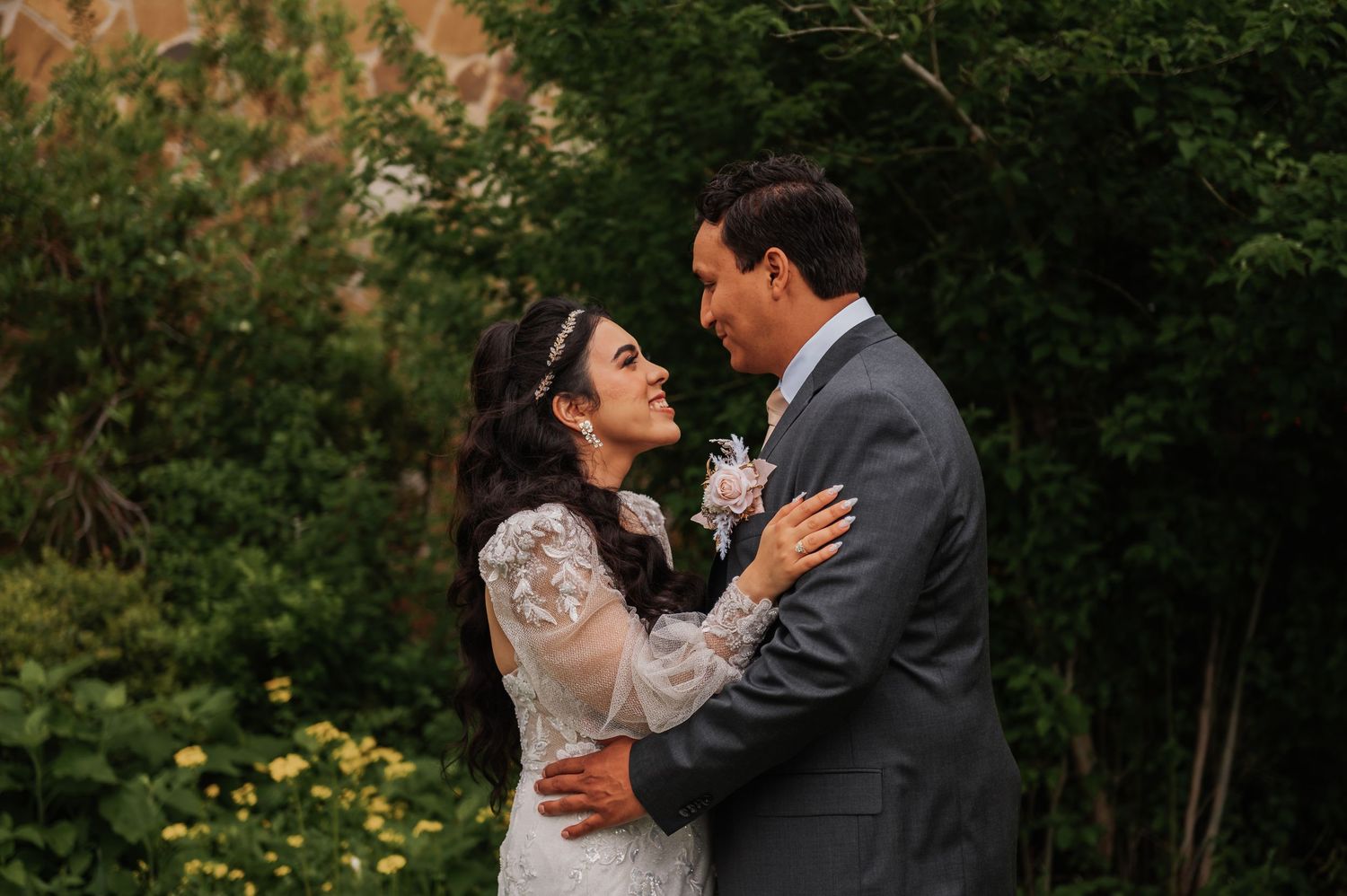 A bride and groom share a romantic moment outdoors, surrounded by lush green foliage, as she wears a lace gown and he a gray suit.