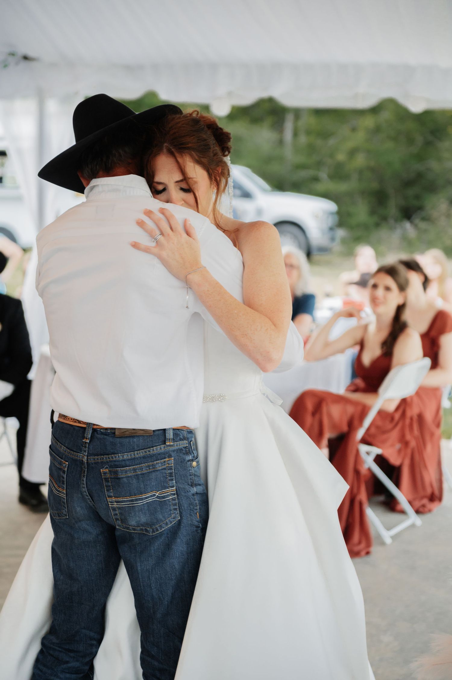 Bride and father share an emotional first dance embrace under a white tent at a rustic wedding reception.