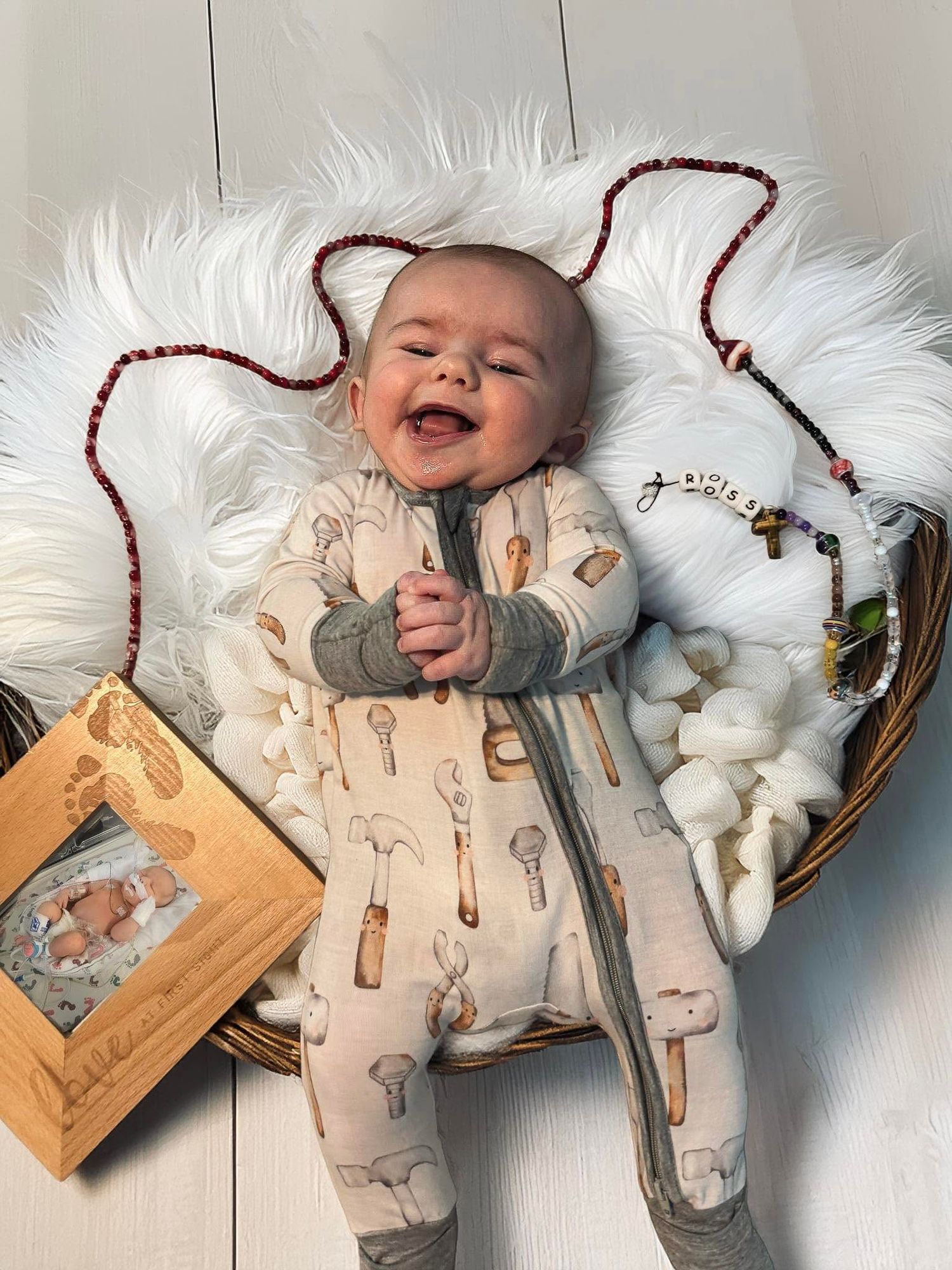 Smiling baby in a tool-print onesie lying in a wicker basket with white fur, rosary beads, and a wooden keepsake box.