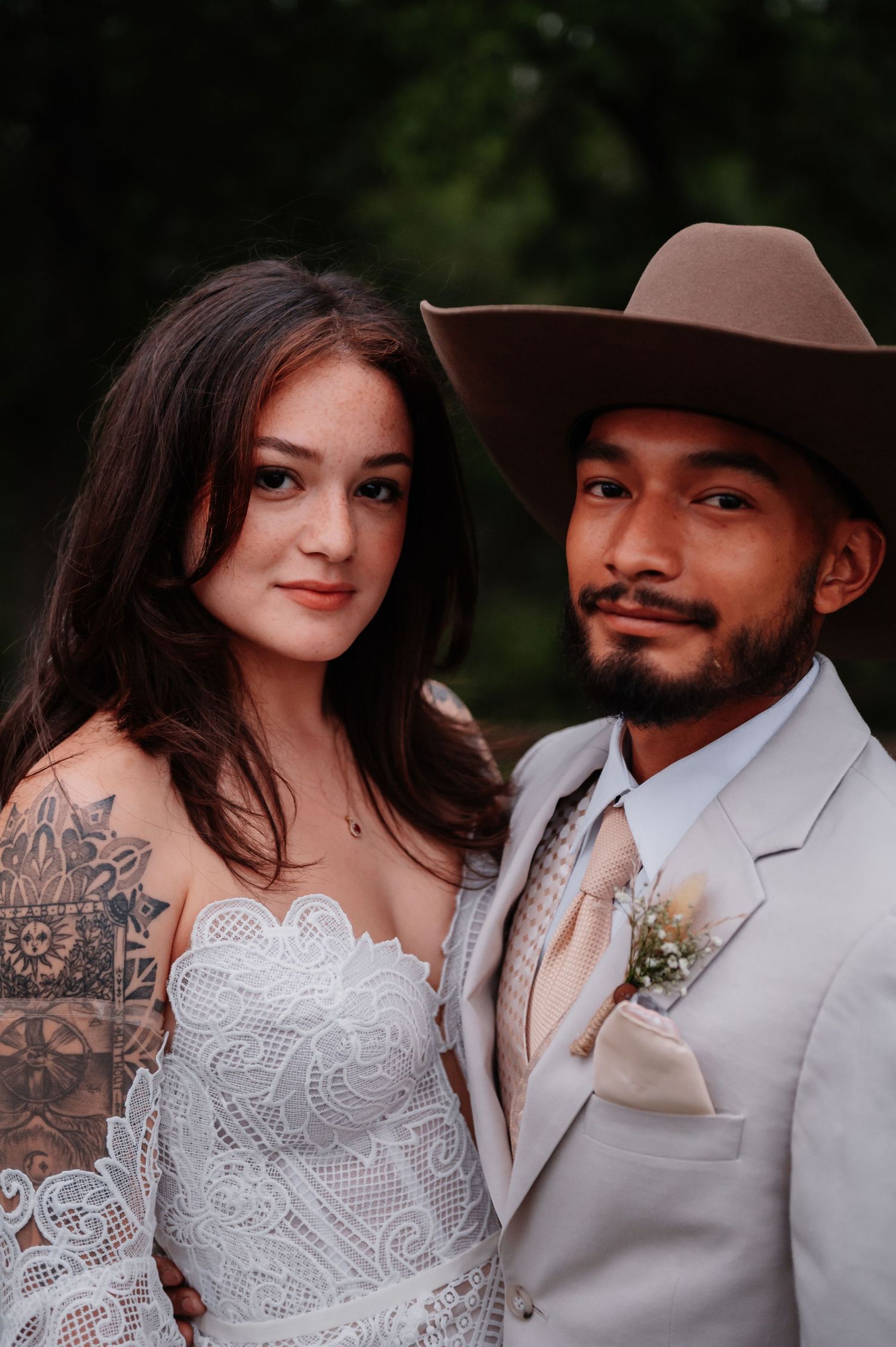 Bride in lace gown with tattoos and groom in tan suit and cowboy hat pose together at outdoor wedding.