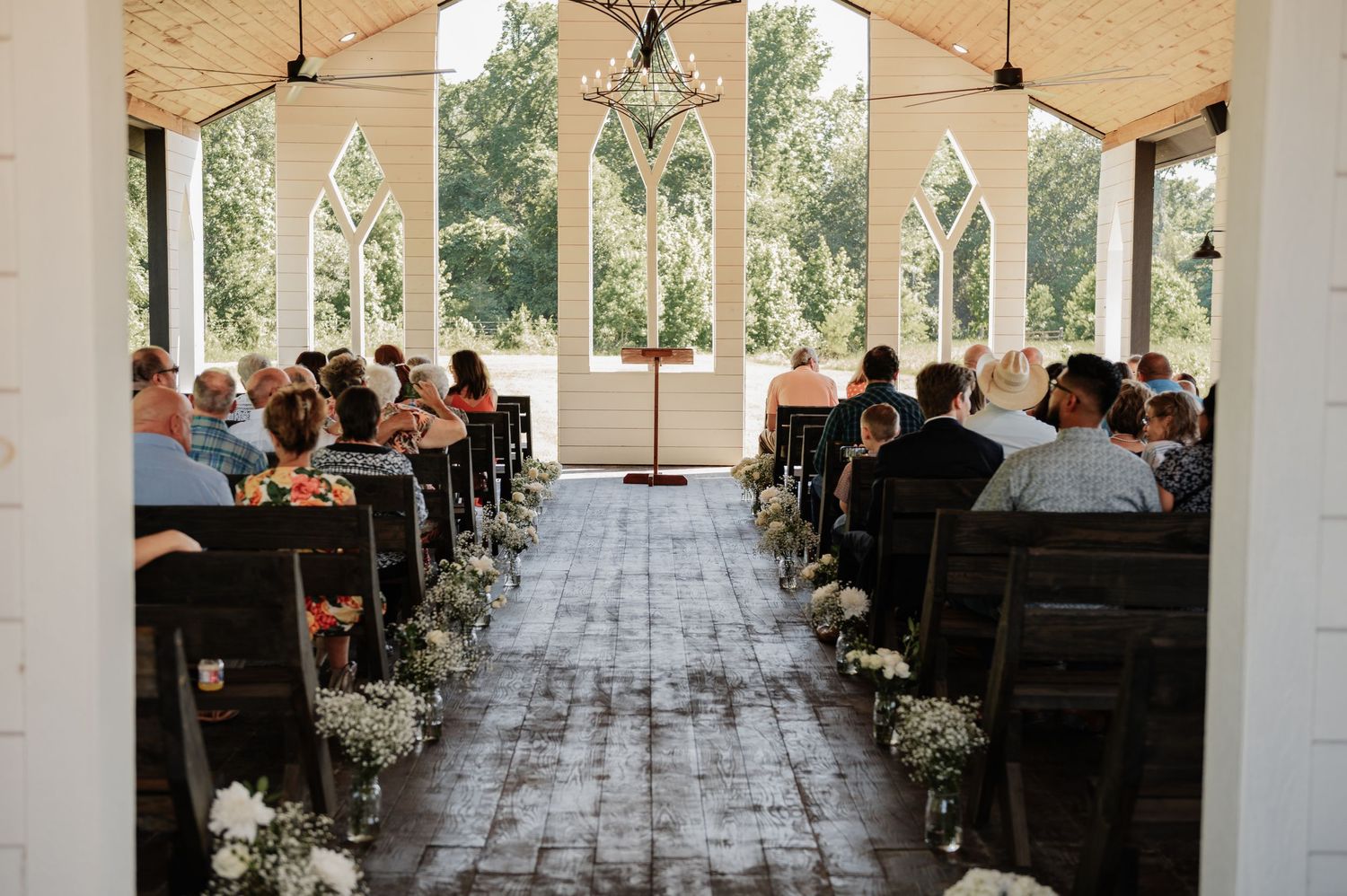 Rustic barn wedding ceremony with wooden pews, floral aisle decor, and Gothic arch windows overlooking lush greenery.