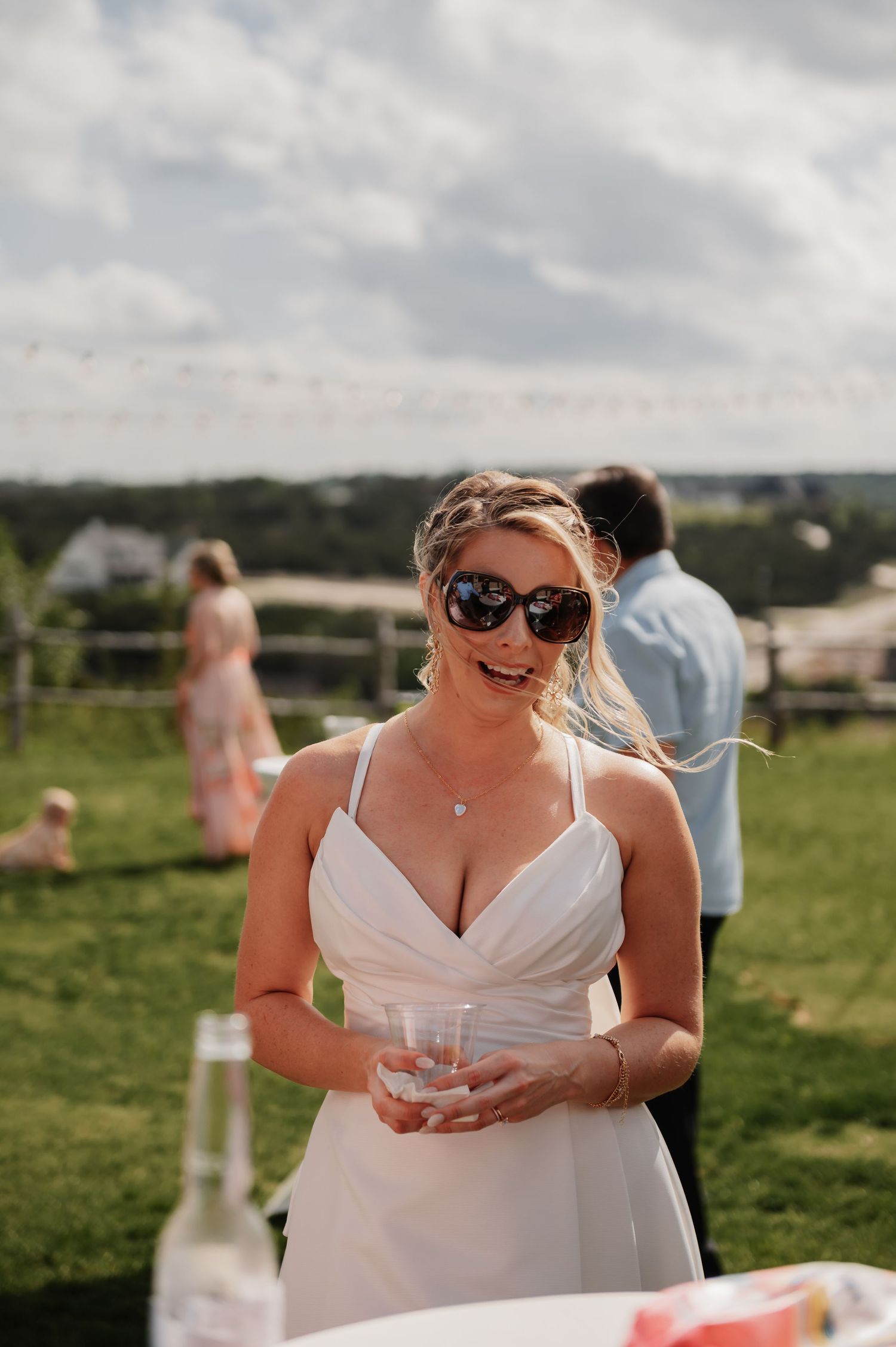 Woman in white dress and sunglasses holding a drink at an outdoor wedding reception on a sunny day.