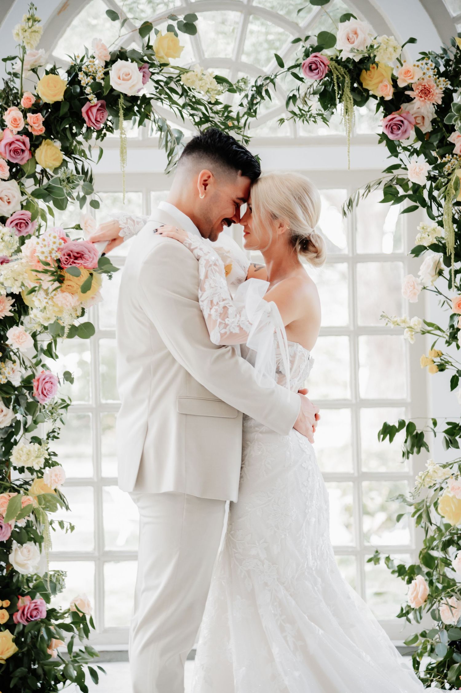 A bride and groom embrace under a floral arch adorned with pink, yellow, and white flowers on their wedding day.