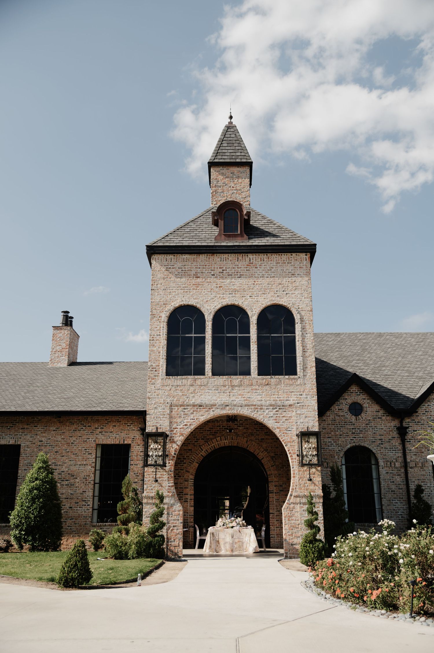 Elegant brick wedding venue with arched entryway, tall tower, and manicured gardens under a blue sky.