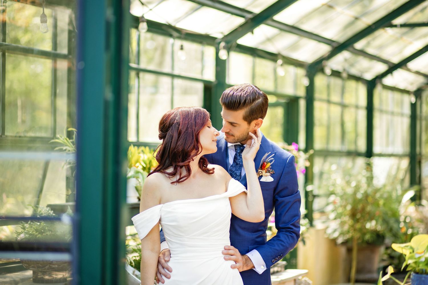 Romantic couple embraces and kisses in a sunlit greenhouse surrounded by lush greenery and glass walls.