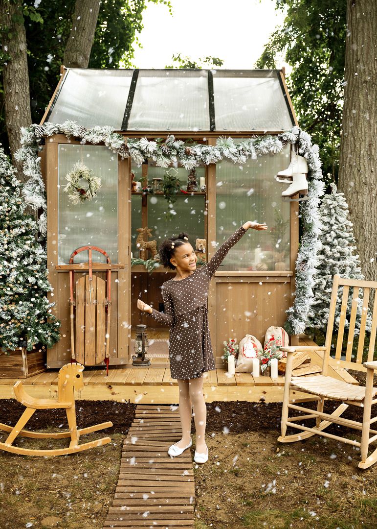 A young girl poses outdoors in falling snow beside a Christmas-decorated greenhouse with rocking chairs and holiday ornaments.