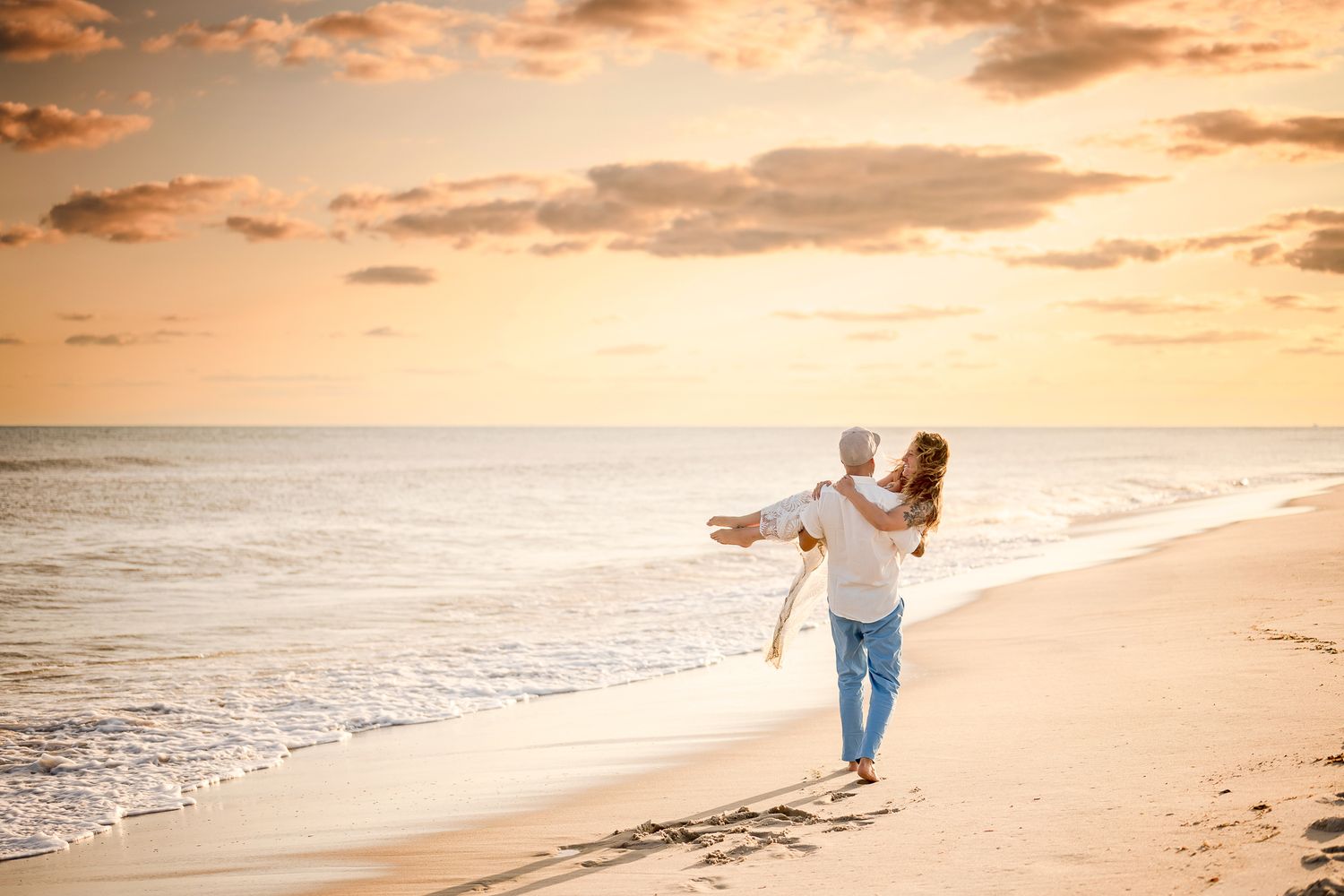 Two silhouettes walking together on a sandy beach at sunset with orange clouds and rolling waves in the background.