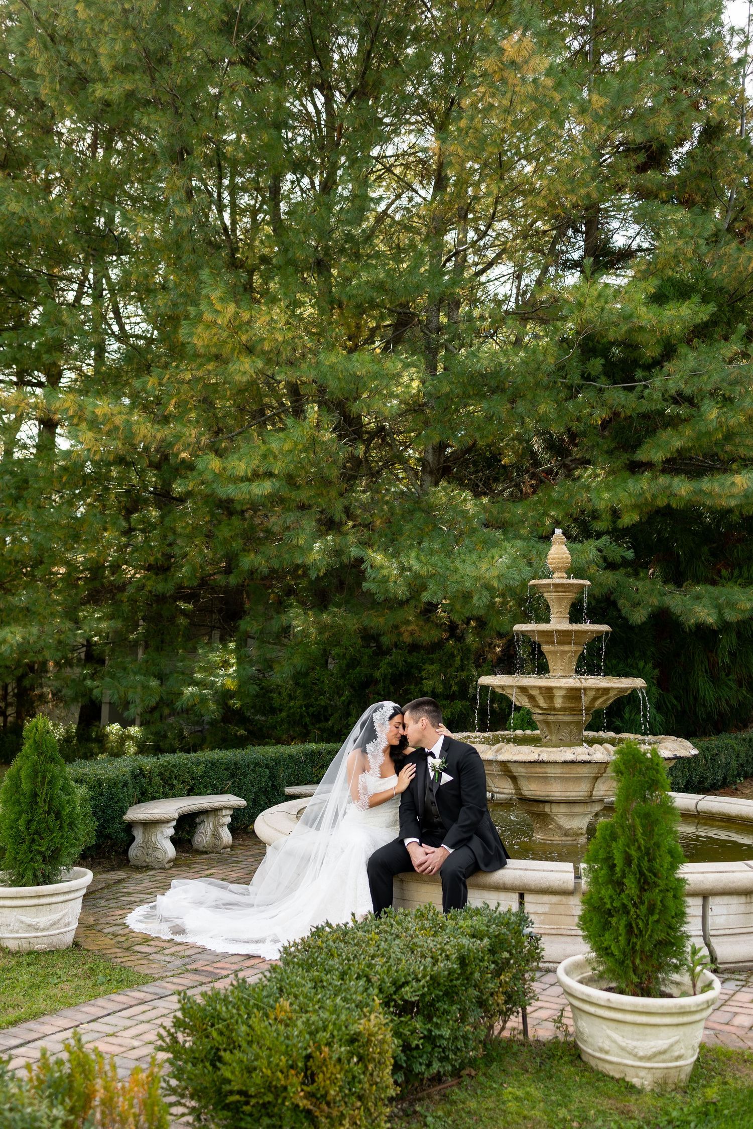 Bride in a white gown and veil sits close with her groom in a black suit on a stone fountain ledge in a lush, tree-lined garden.