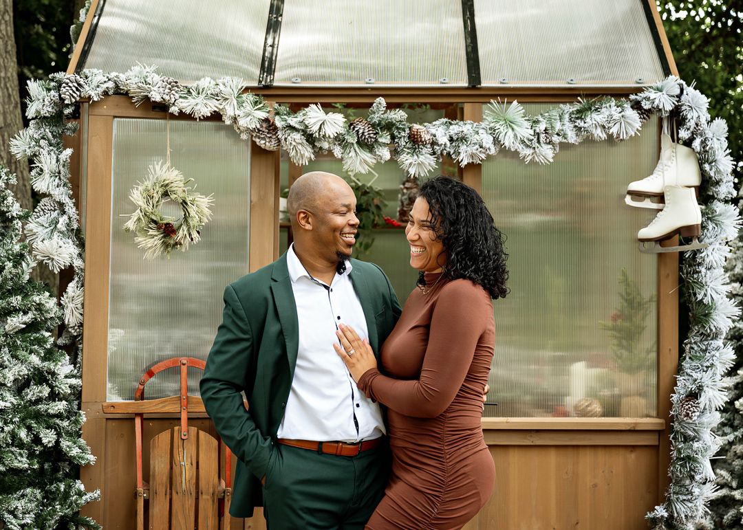 A couple shares a joyful moment in front of a Christmas-decorated greenhouse with garland and ice skates.