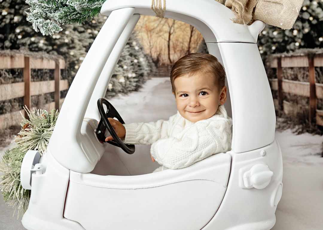 Smiling baby in white outfit sits in a toy car surrounded by snowy trees in a winter photo session.