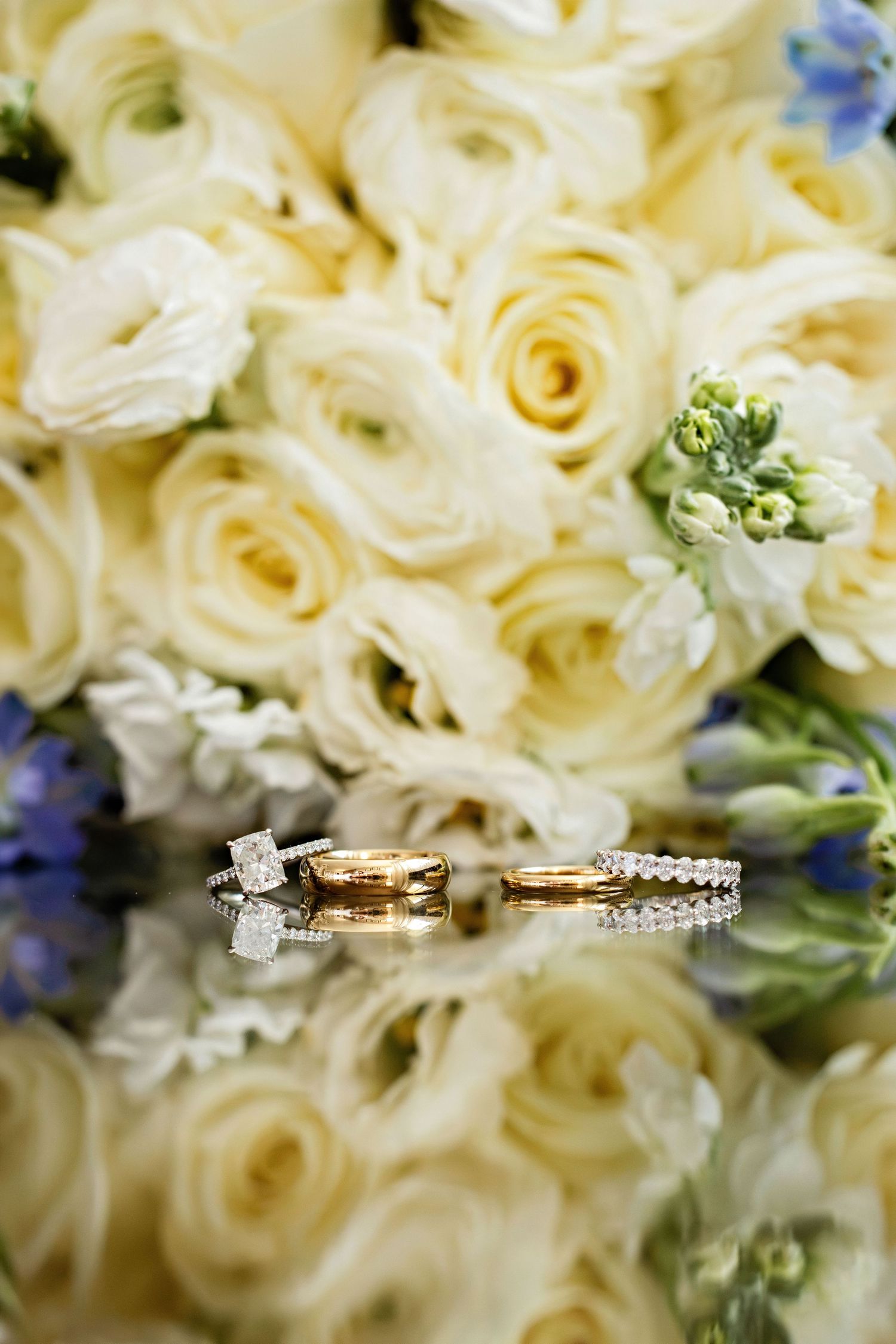 Gold wedding rings with diamonds rest on a reflective surface before a bouquet of white roses and blue flowers.