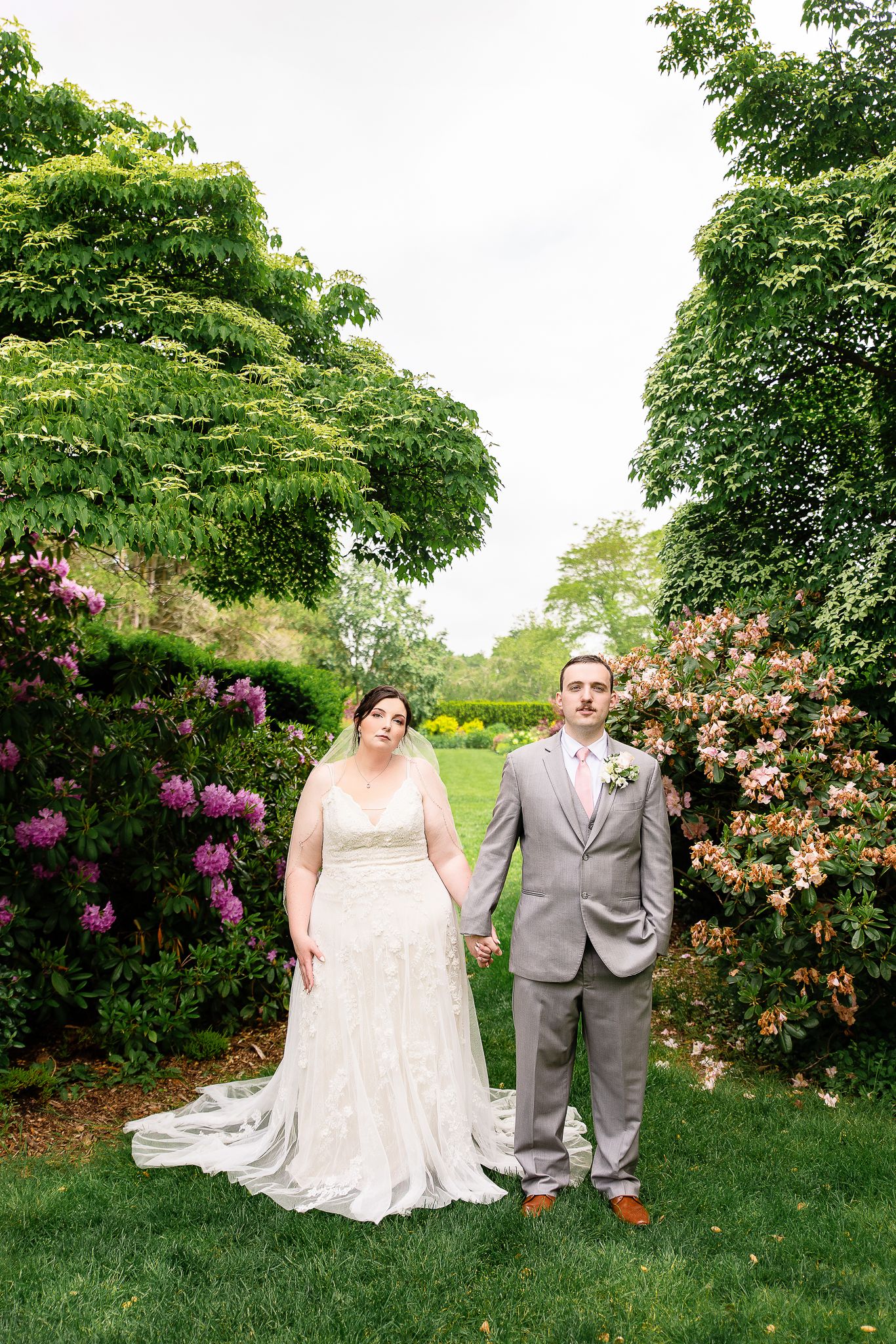 A wedding couple stands in a lush garden setting with blooming rhododendrons and green trees creating a romantic backdrop.