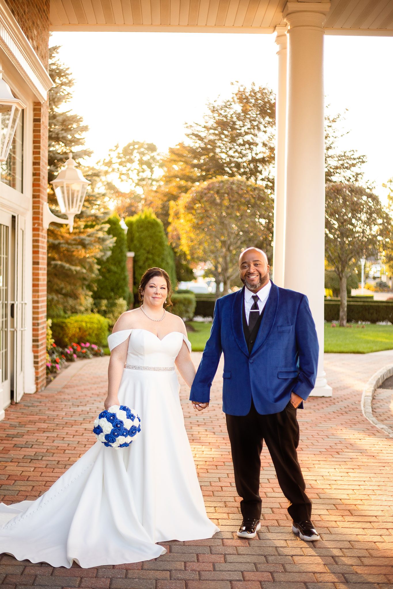 A couple stands together on a brick walkway at golden hour, the bride holding a blue bouquet and wearing a white gown.